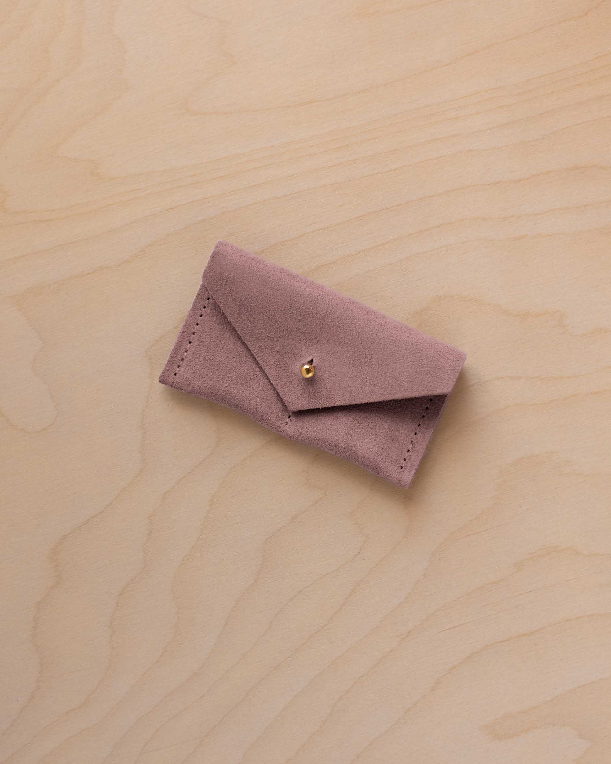 A small dusty mauve coloured suede jewellery pouch with a gold coloured fastening, resting on a light wood grain surface.