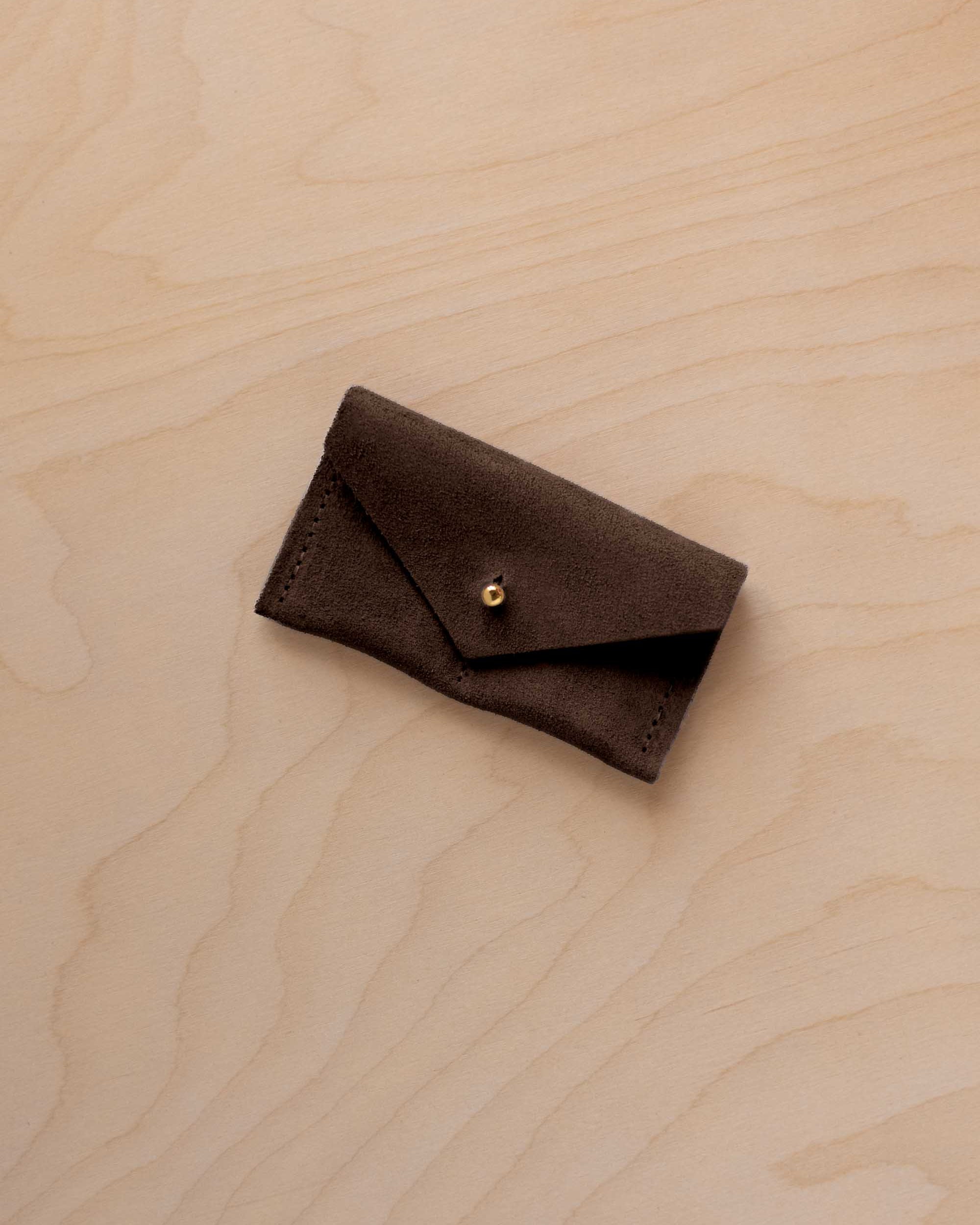 A small espresso brown coloured suede jewellery pouch with a gold button, resting on a light wood grain surface.