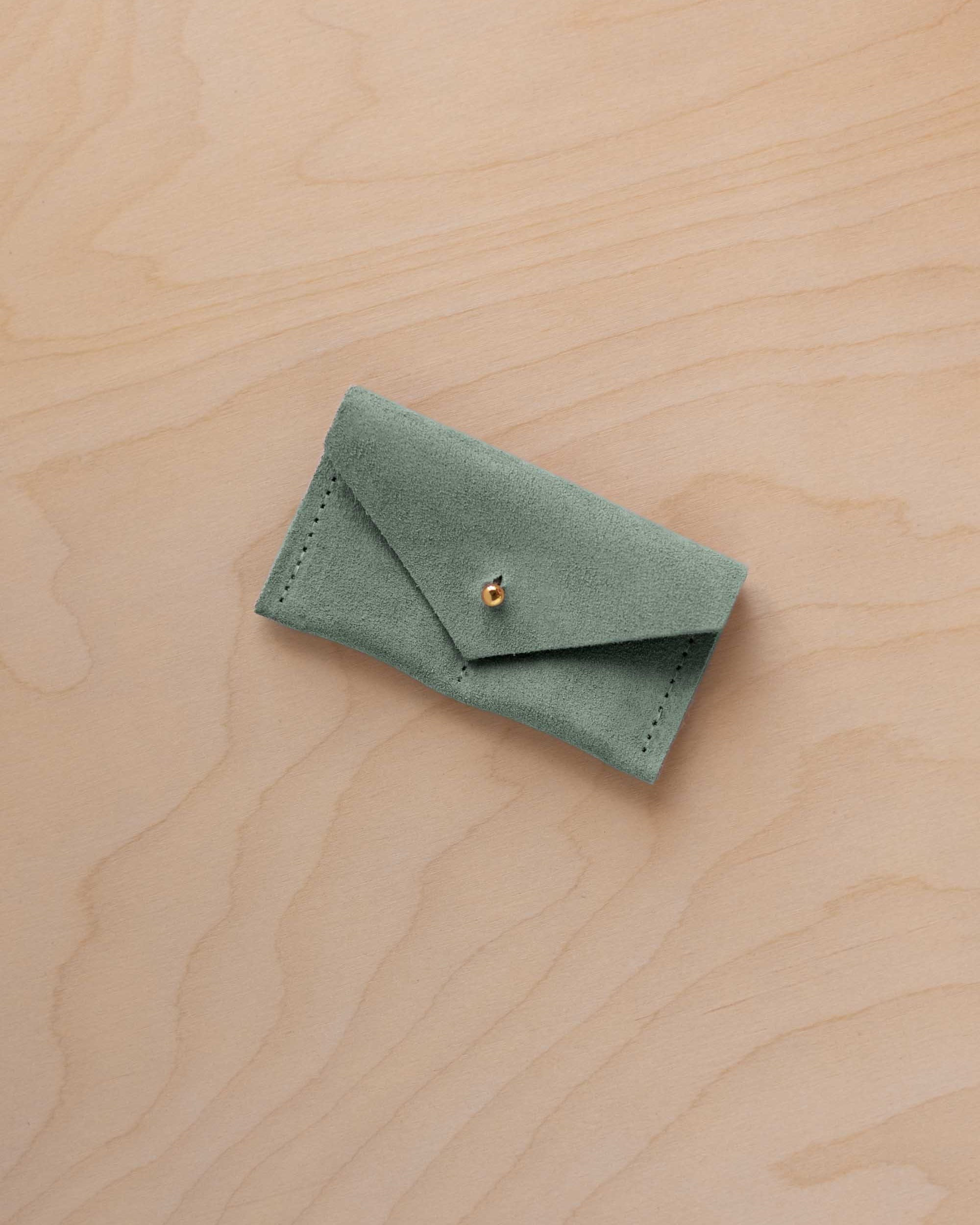A small eucalyptus green coloured suede jewellery pouch with a gold coloured fastening, resting on a light wood grain surface.