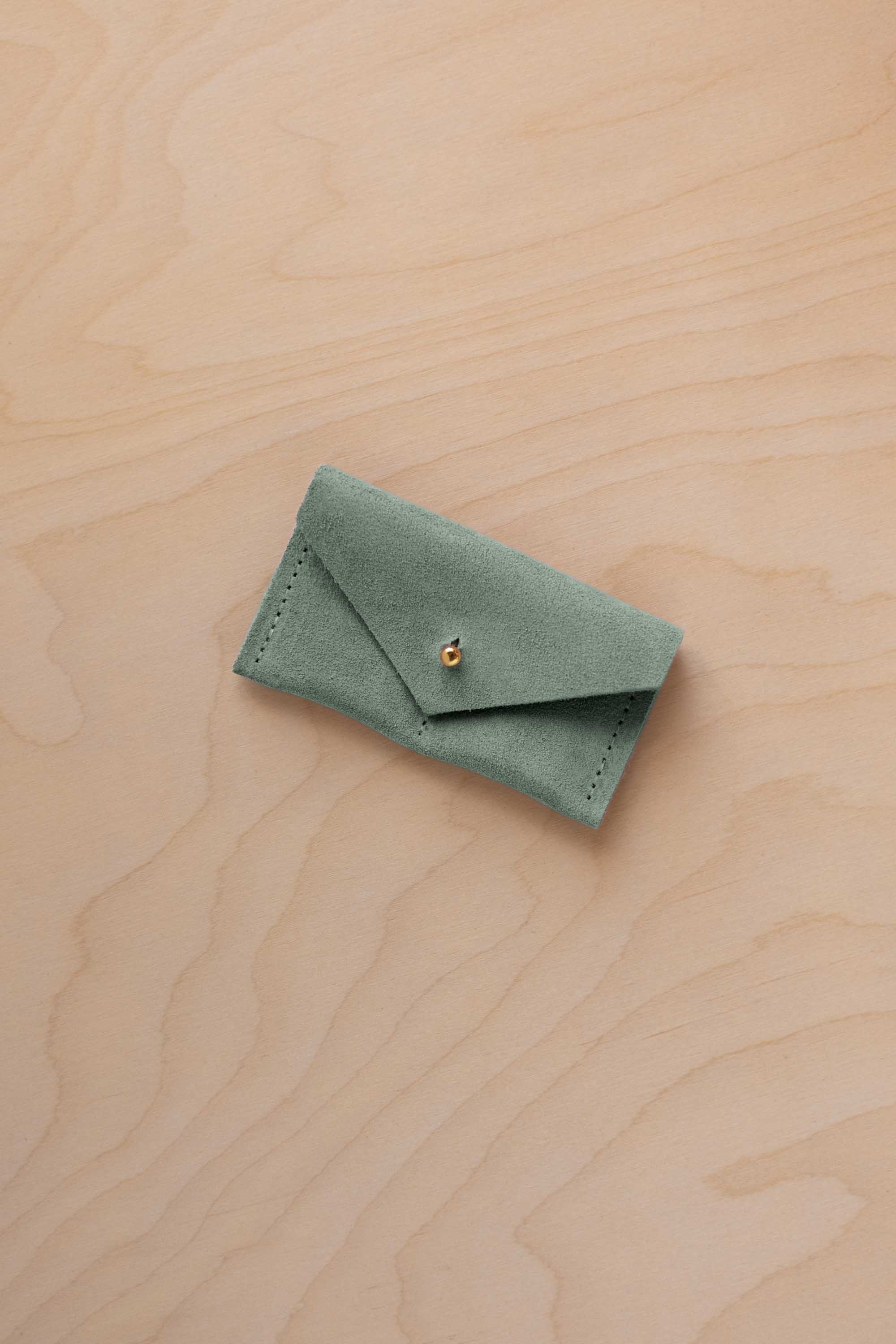 A small eucalyptus green coloured suede jewellery pouch with a gold coloured fastening, resting on a light wood grain surface.