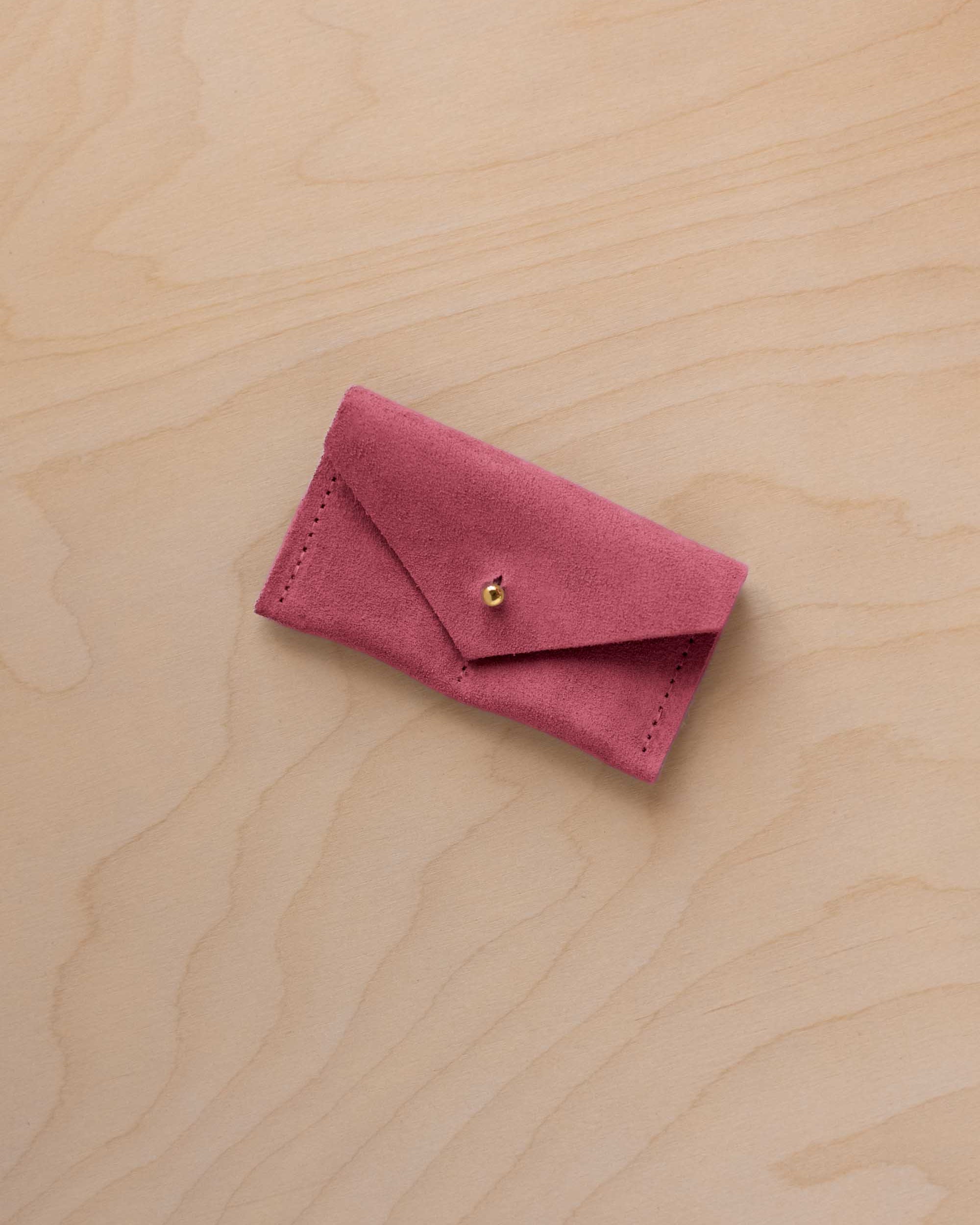 A small faded rose coloured suede jewellery pouch with a gold coloured fastening, resting on a light wood grain surface.