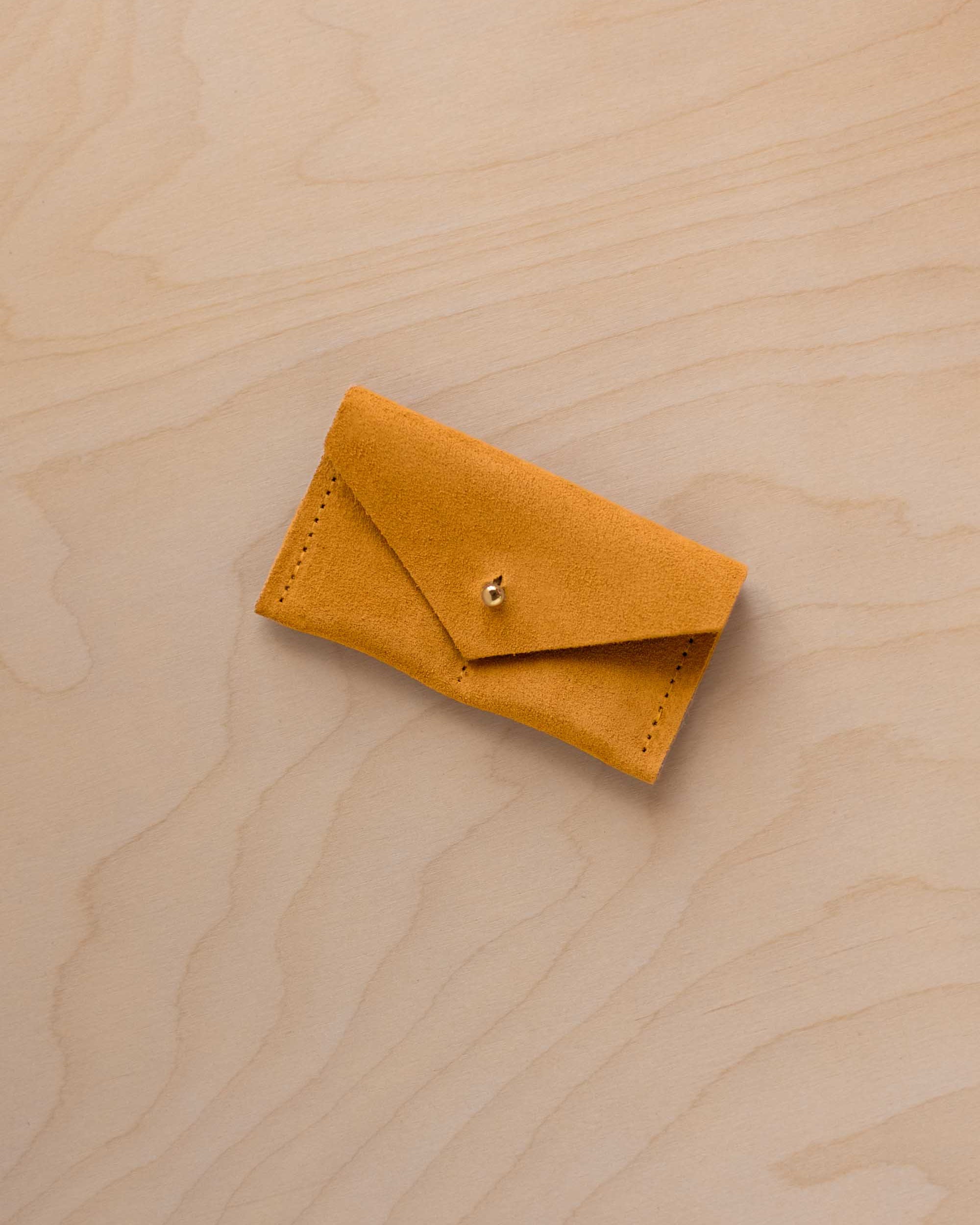 A small golden ochre coloured suede jewellery pouch with a gold coloured fastening, resting on a light wood grain surface.