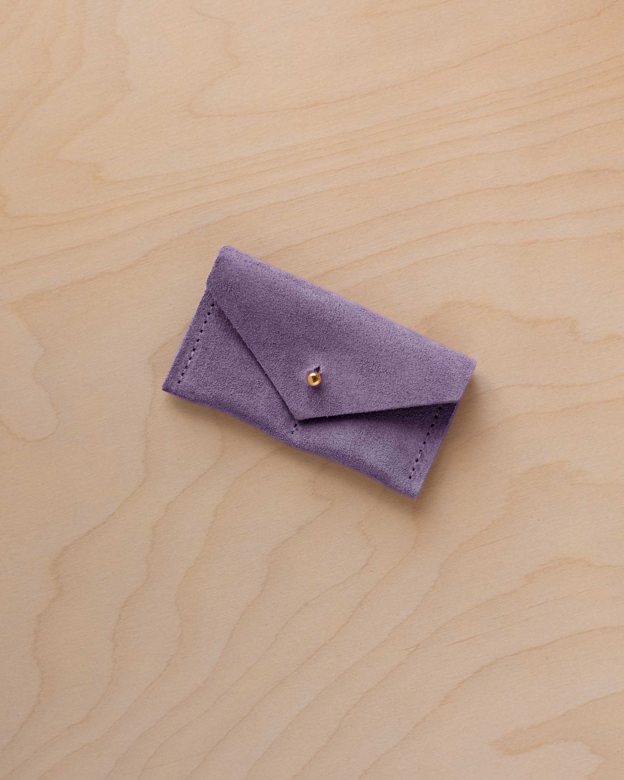 A small lavender coloured suede jewellery pouch with a gold coloured fastening, resting on a light wood grain surface.
