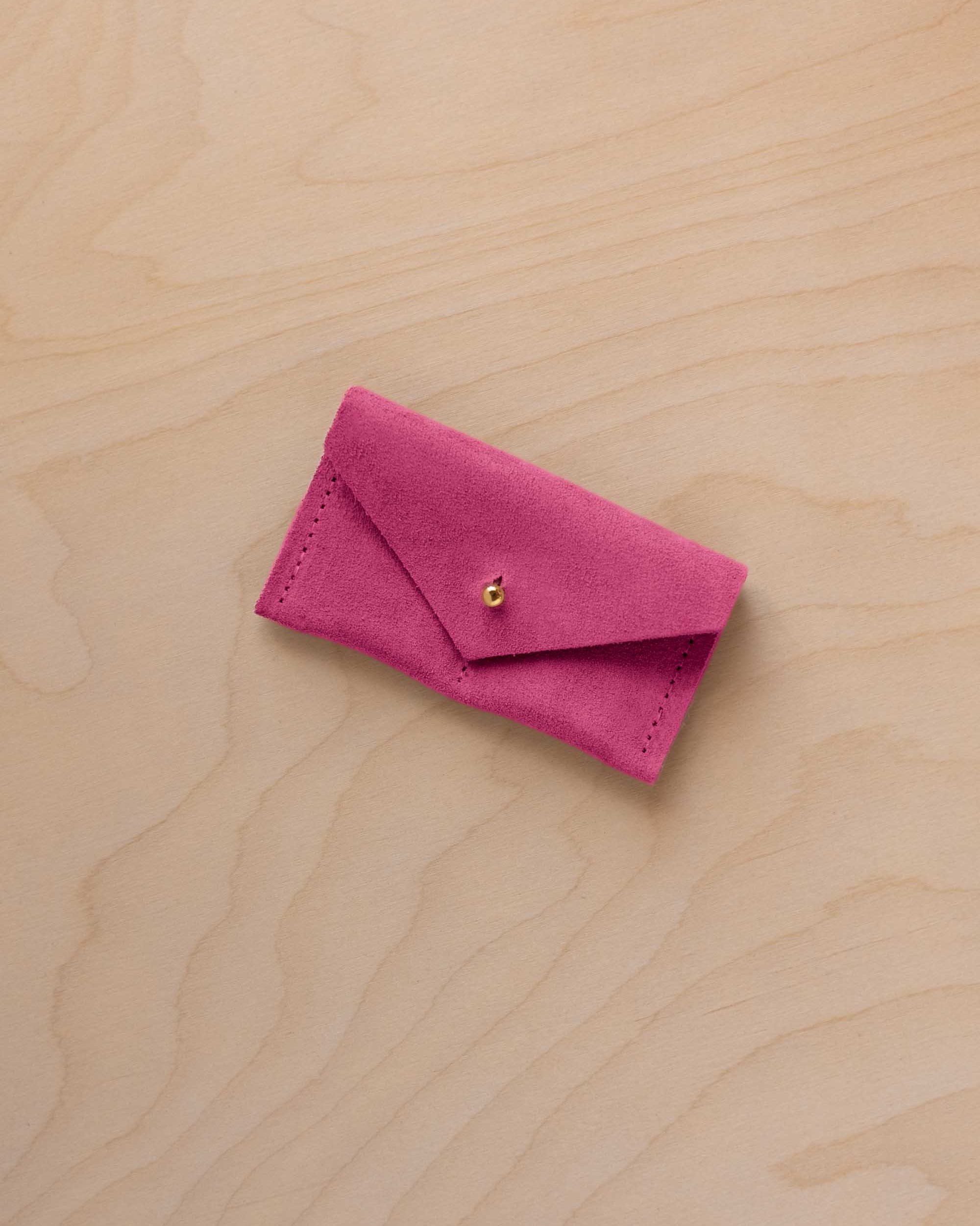 A small raspberry coloured suede jewellery pouch with a gold coloured fastening, resting on a light wood grain surface.