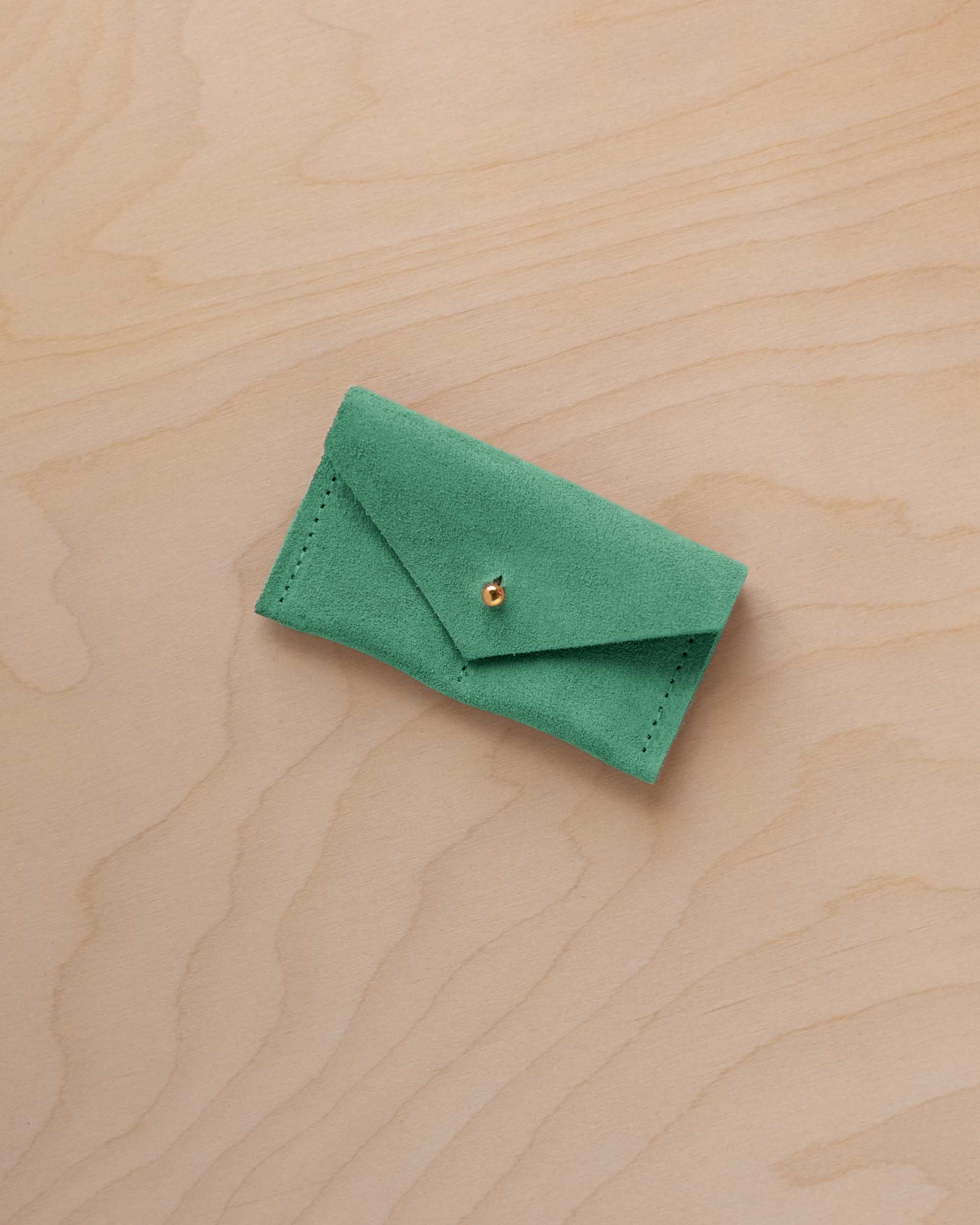 A small seafoam green coloured suede jewellery pouch with a gold coloured fastening, resting on a light wood grain surface.