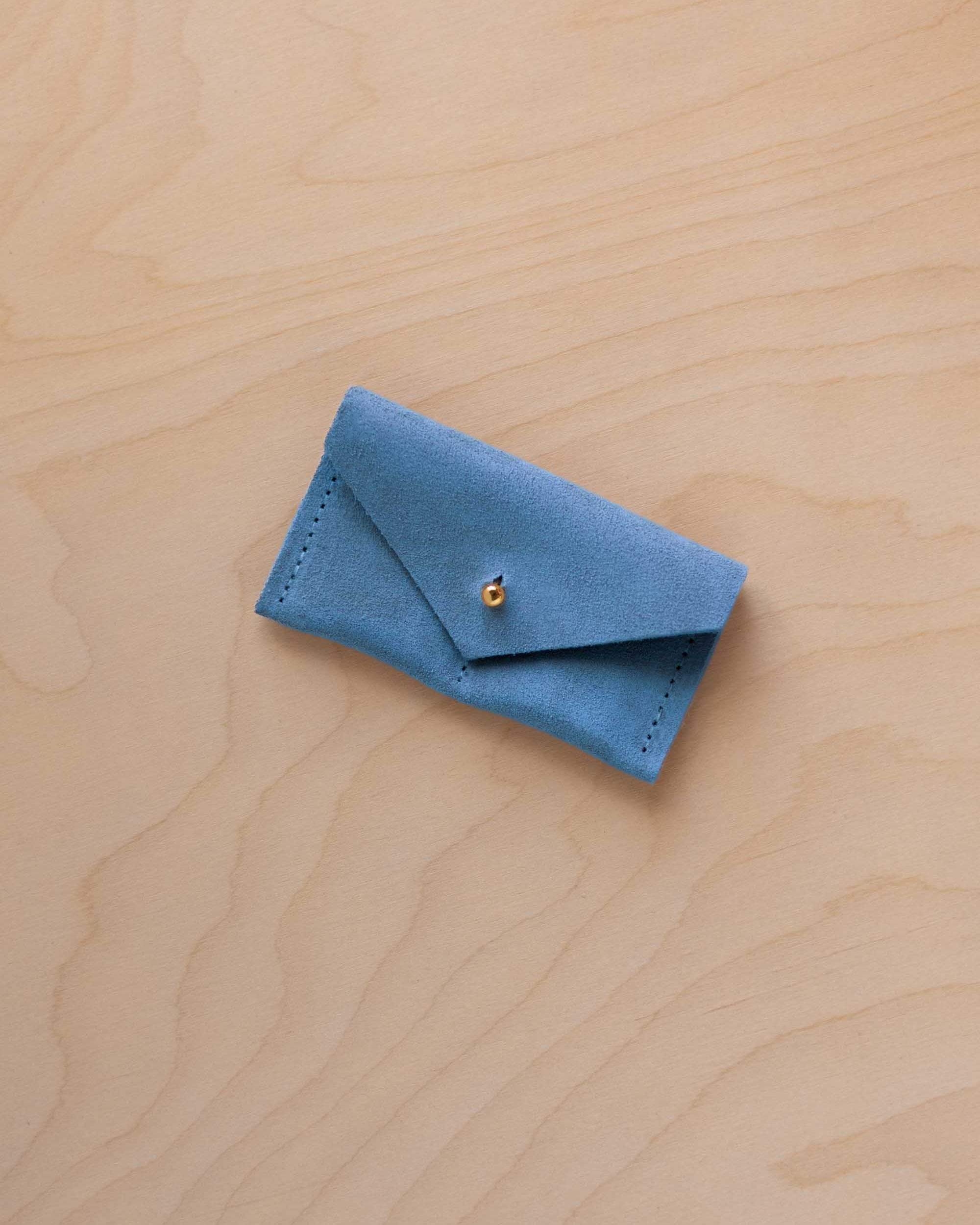 A small sky blue coloured suede jewellery pouch with a gold coloured fastening, resting on a light wood grain surface.