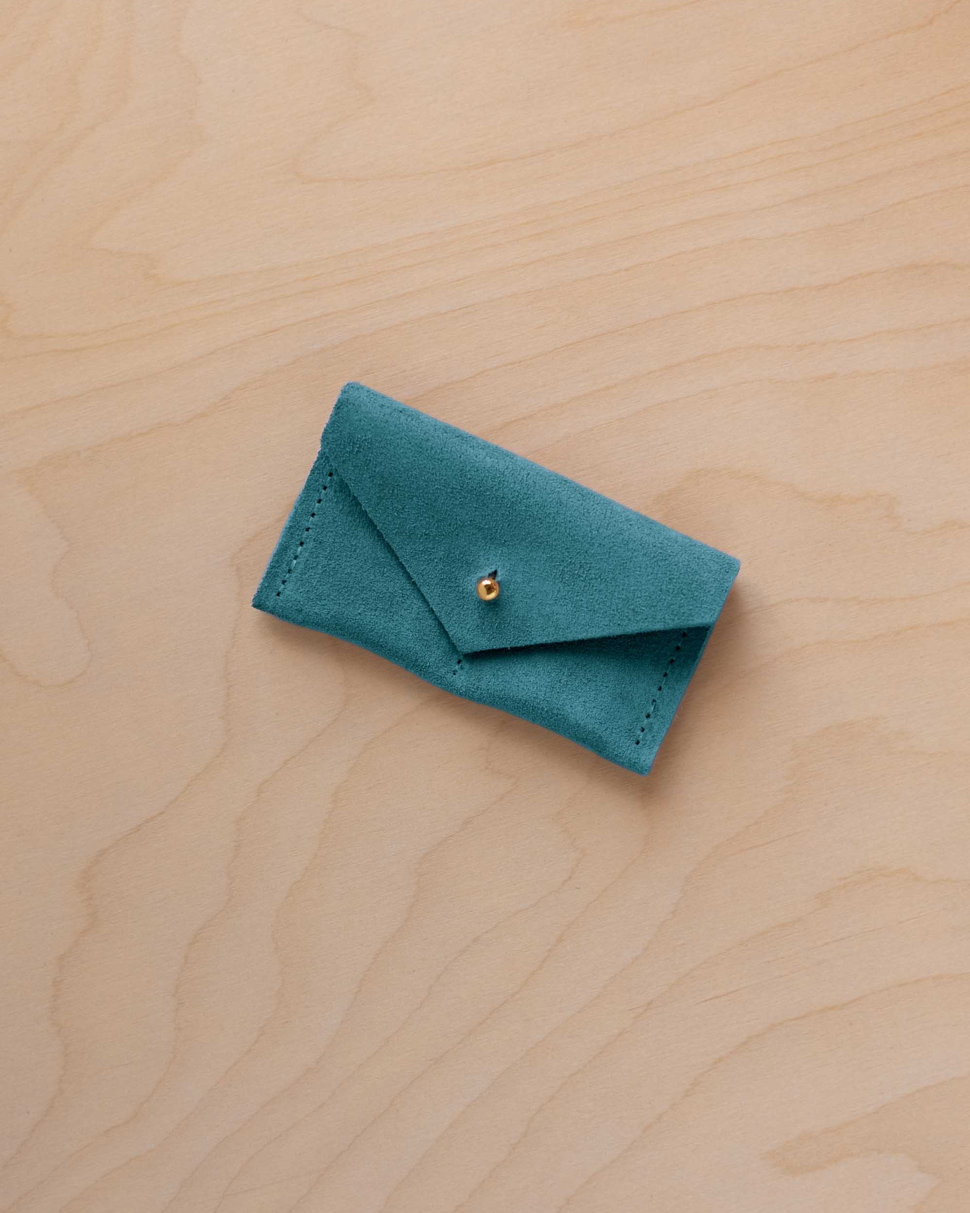 A small marine teal coloured suede jewellery pouch with a gold coloured fastening, resting on a light wood grain surface.