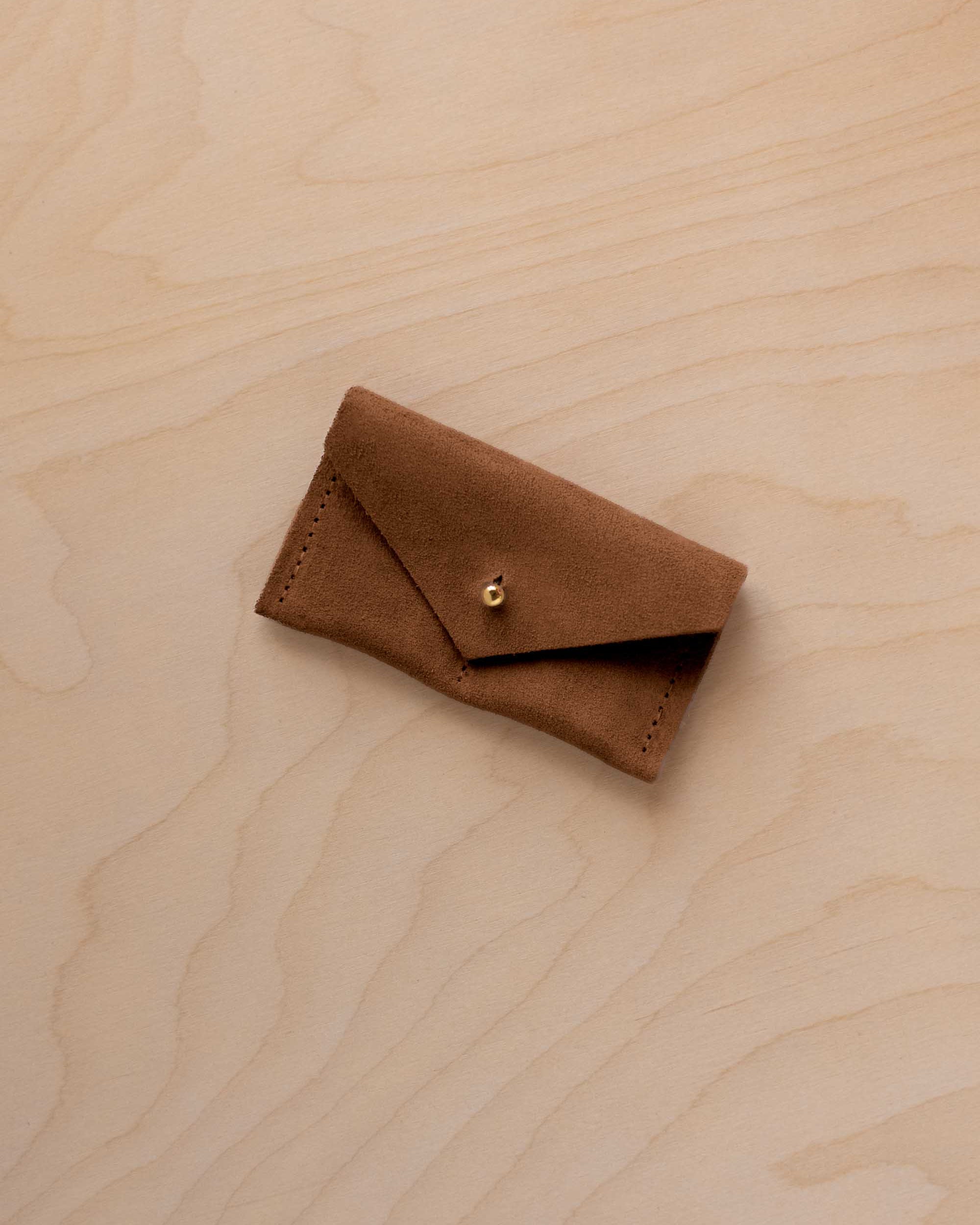 A small mocha brown coloured suede jewellery pouch with a gold coloured fastening, resting on a light wood grain surface.