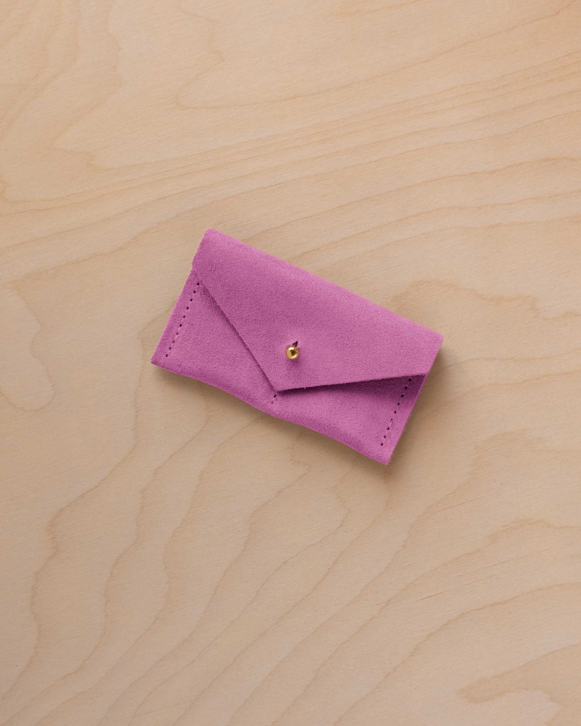A small peony pink coloured suede jewellery pouch with a gold coloured fastening, resting on a light wood grain surface.