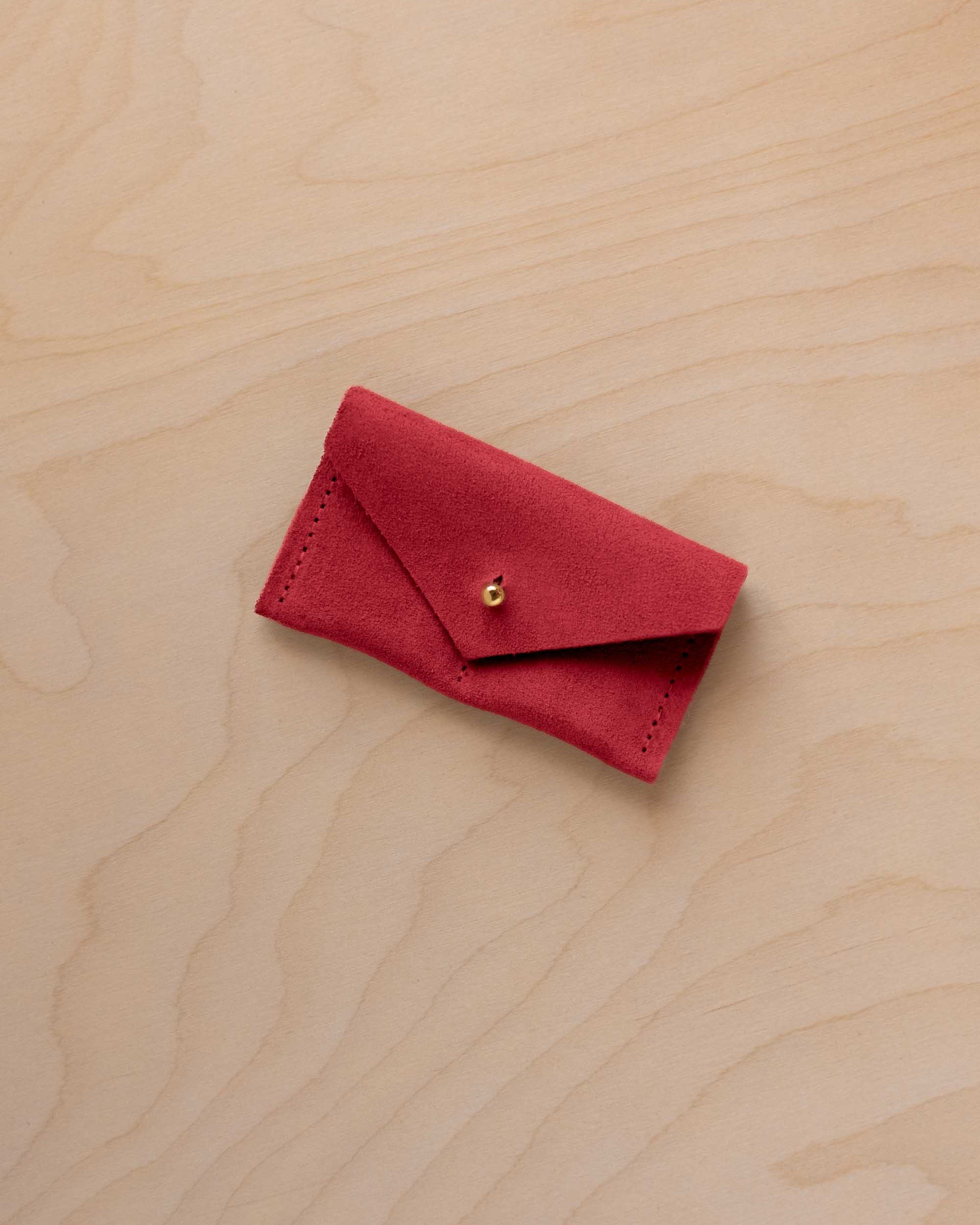 A small scarlet red coloured suede jewellery pouch with a gold coloured fastening, resting on a light wood grain surface.