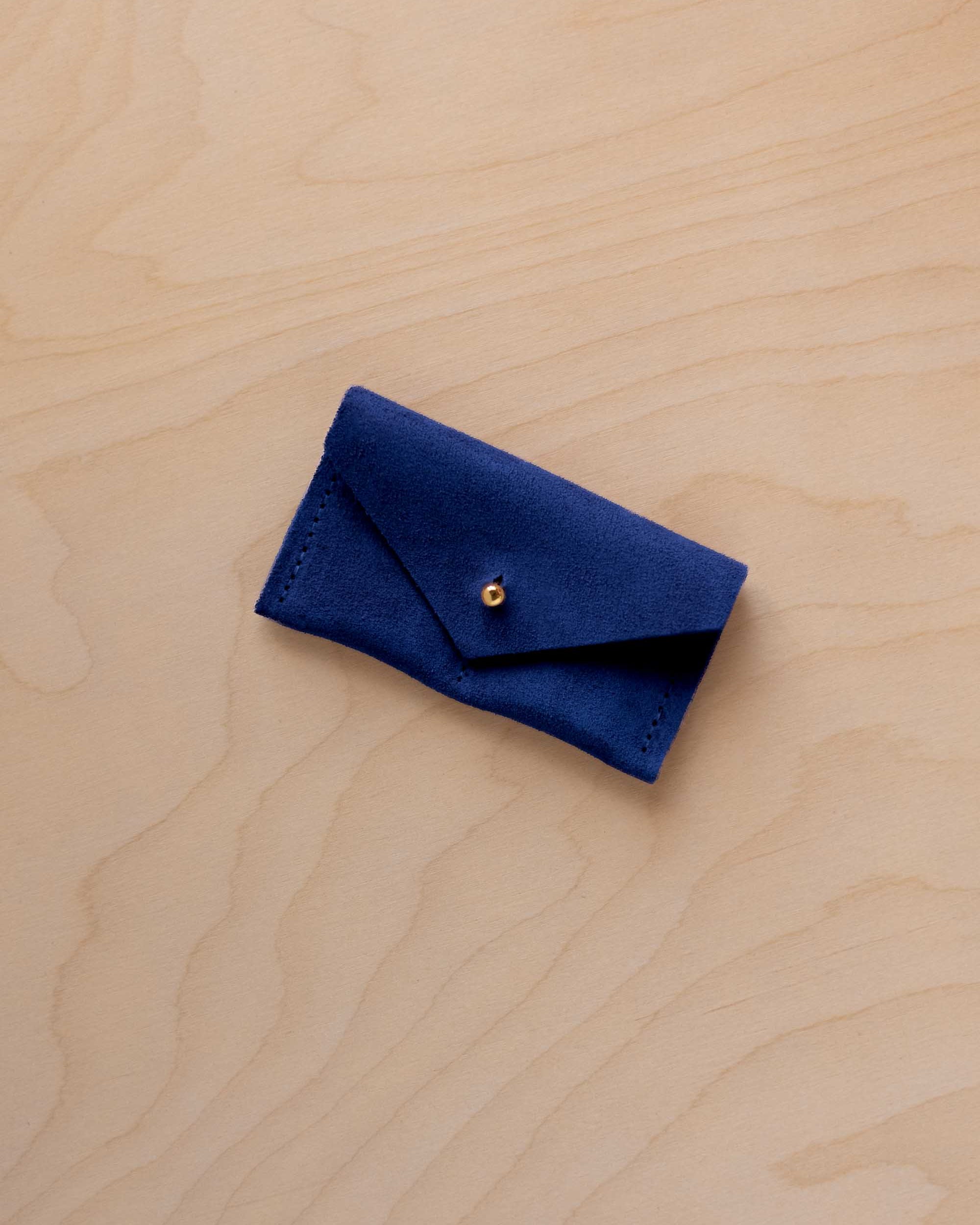 A small ultramarine blue coloured suede jewellery pouch with a gold coloured fastening, resting on a light wood grain surface.