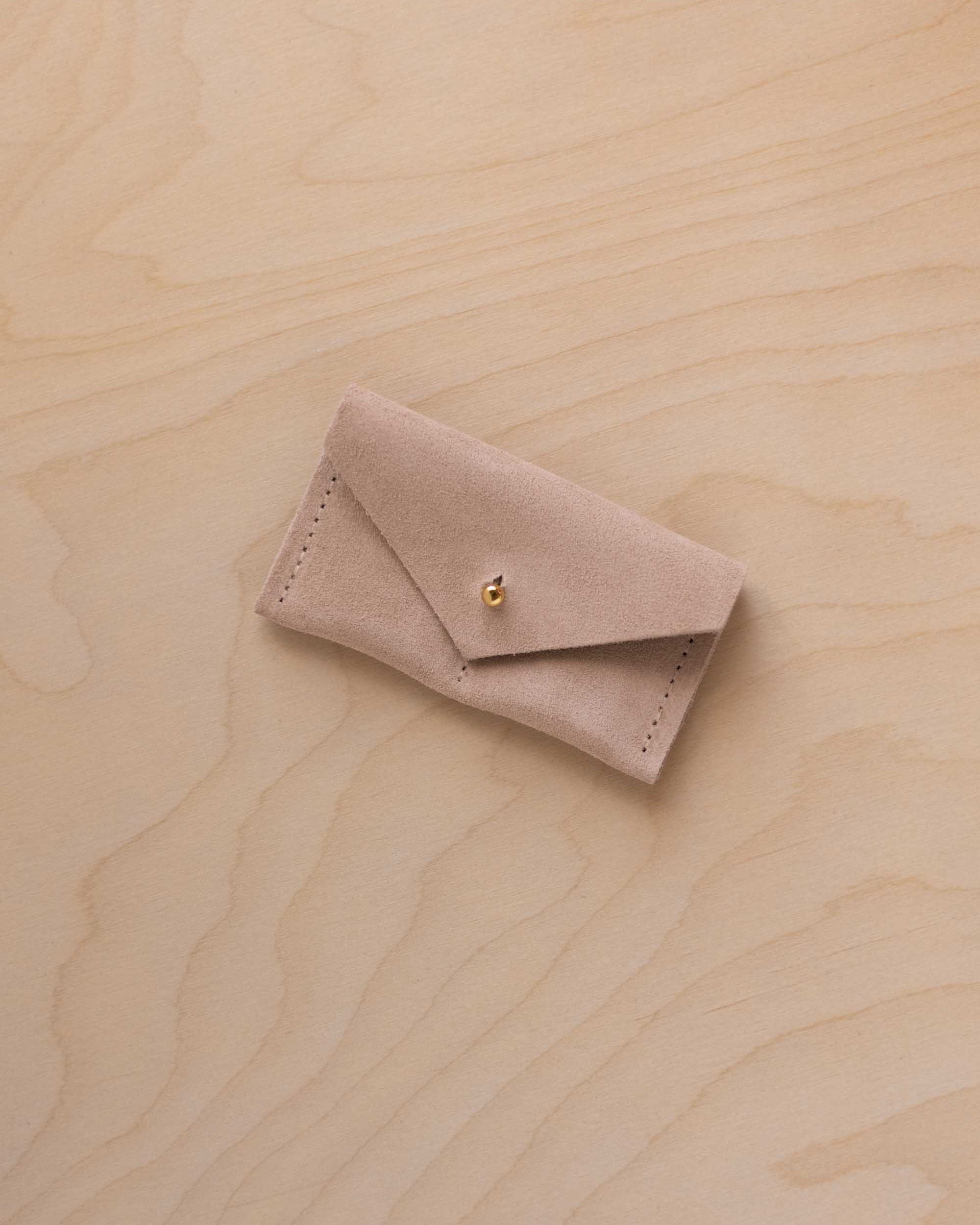 A small warm sand coloured suede jewellery pouch with a gold coloured fastening, resting on a light wood grain surface.