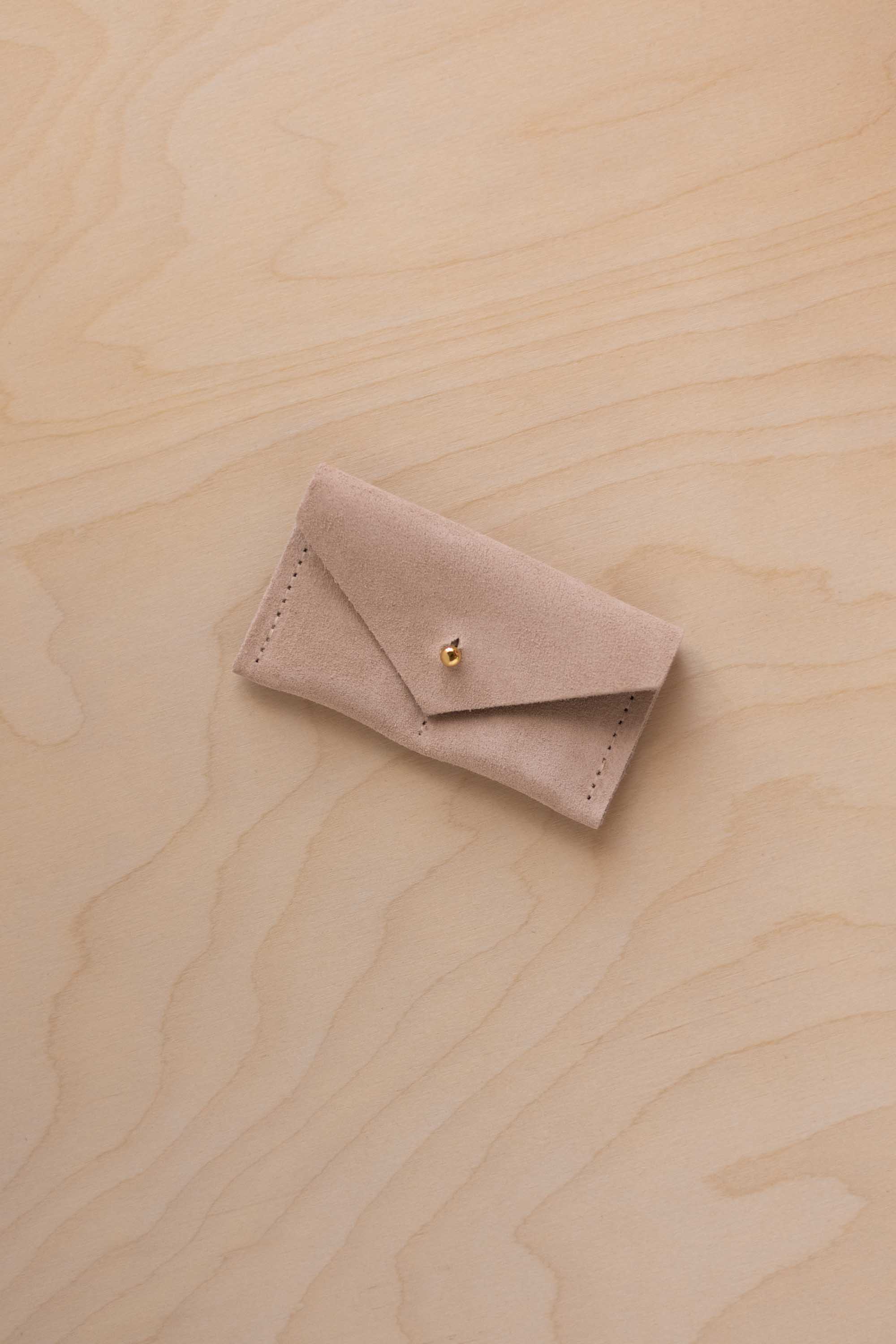 A small warm sand coloured suede jewellery pouch with a gold coloured fastening, resting on a light wood grain surface.