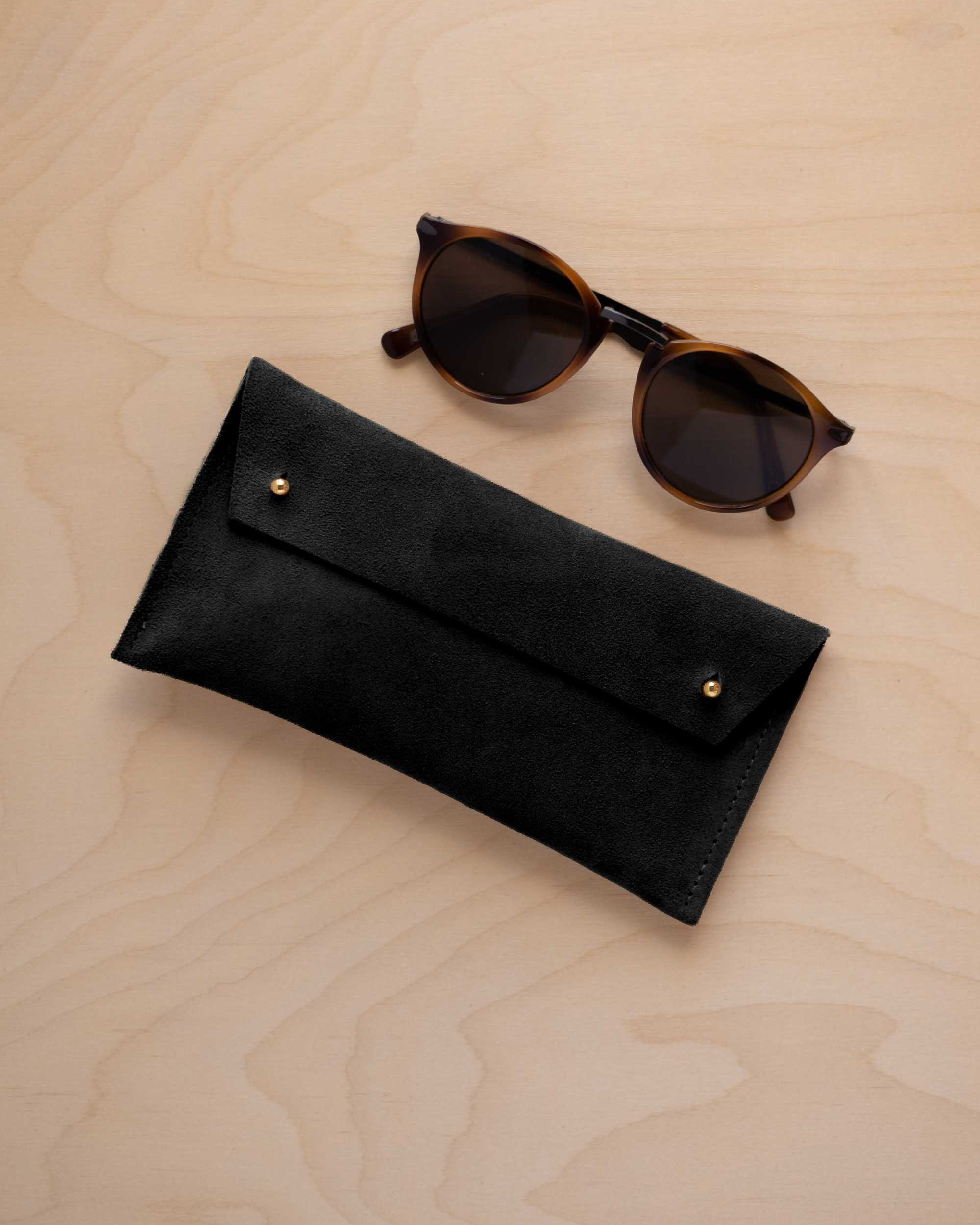 A pair of dark brown tortoiseshell sunglasses with round frames, resting on a light beige wood-grain surface next to a black suede glasses case with gold coloured buttons.