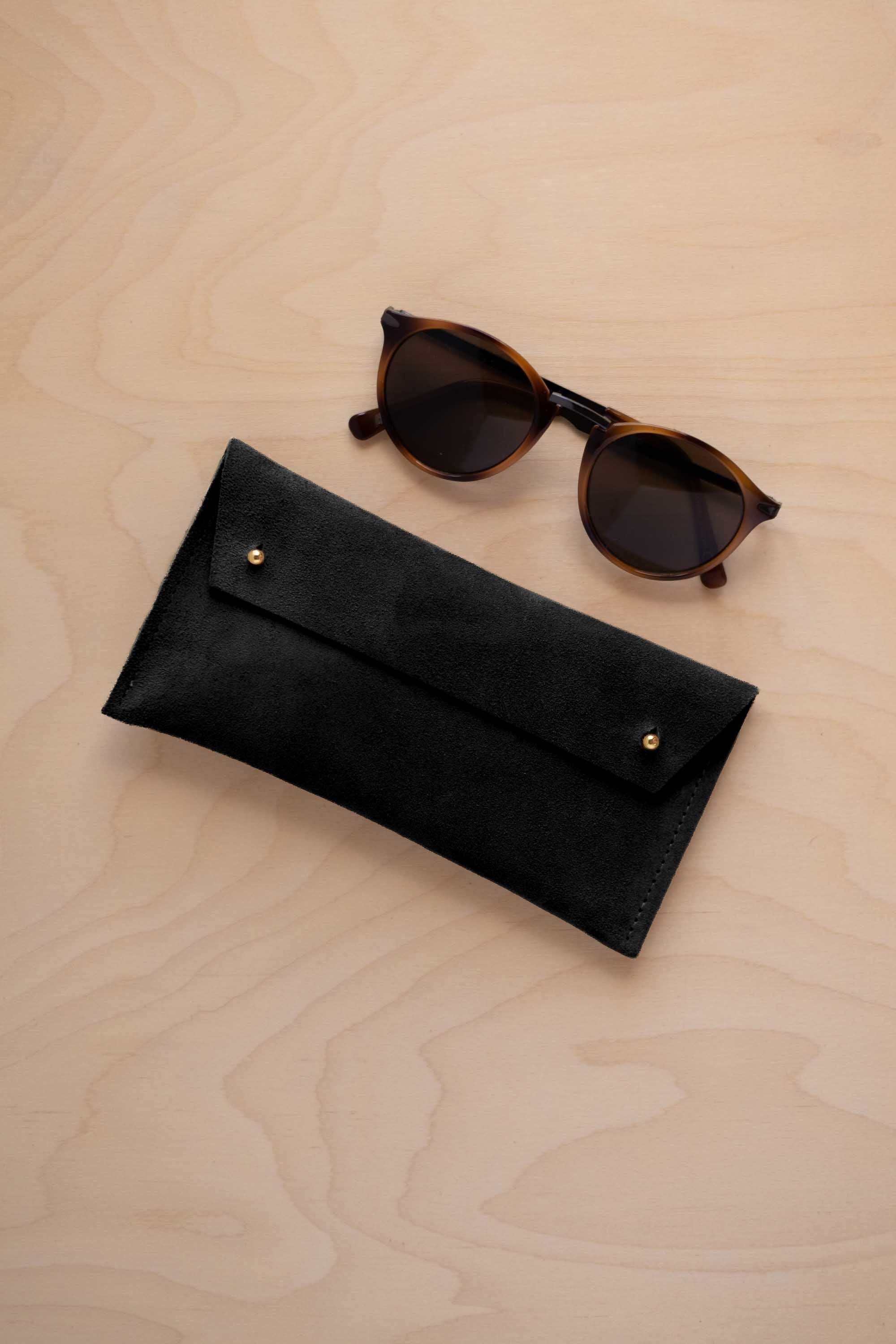 A pair of dark brown tortoiseshell sunglasses with round frames, resting on a light beige wood-grain surface next to a black suede glasses case with gold coloured buttons.