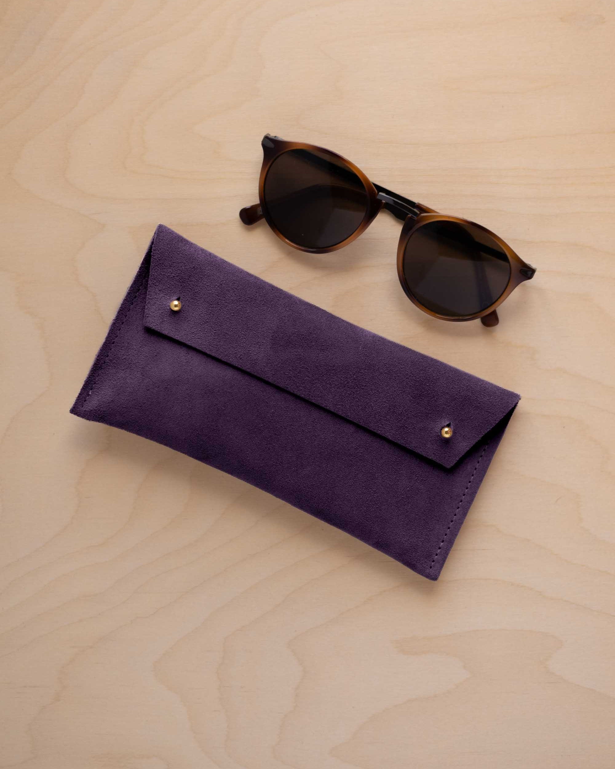 A pair of dark brown tortoiseshell sunglasses with round frames, resting on a light beige wood-grain surface next to a blackcurrant dark purple coloured suede glasses case with gold coloured buttons.