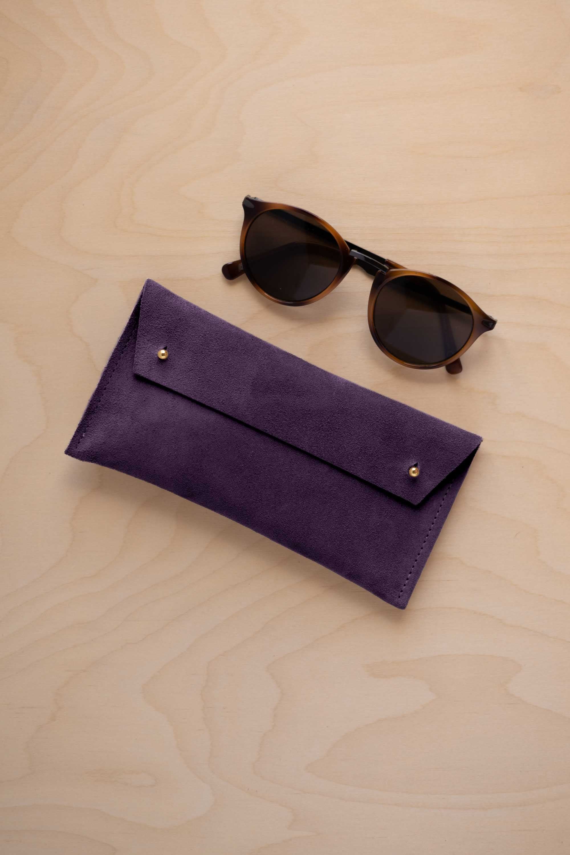 A pair of dark brown tortoiseshell sunglasses with round frames, resting on a light beige wood-grain surface next to a blackcurrant dark purple coloured suede glasses case with gold coloured buttons.