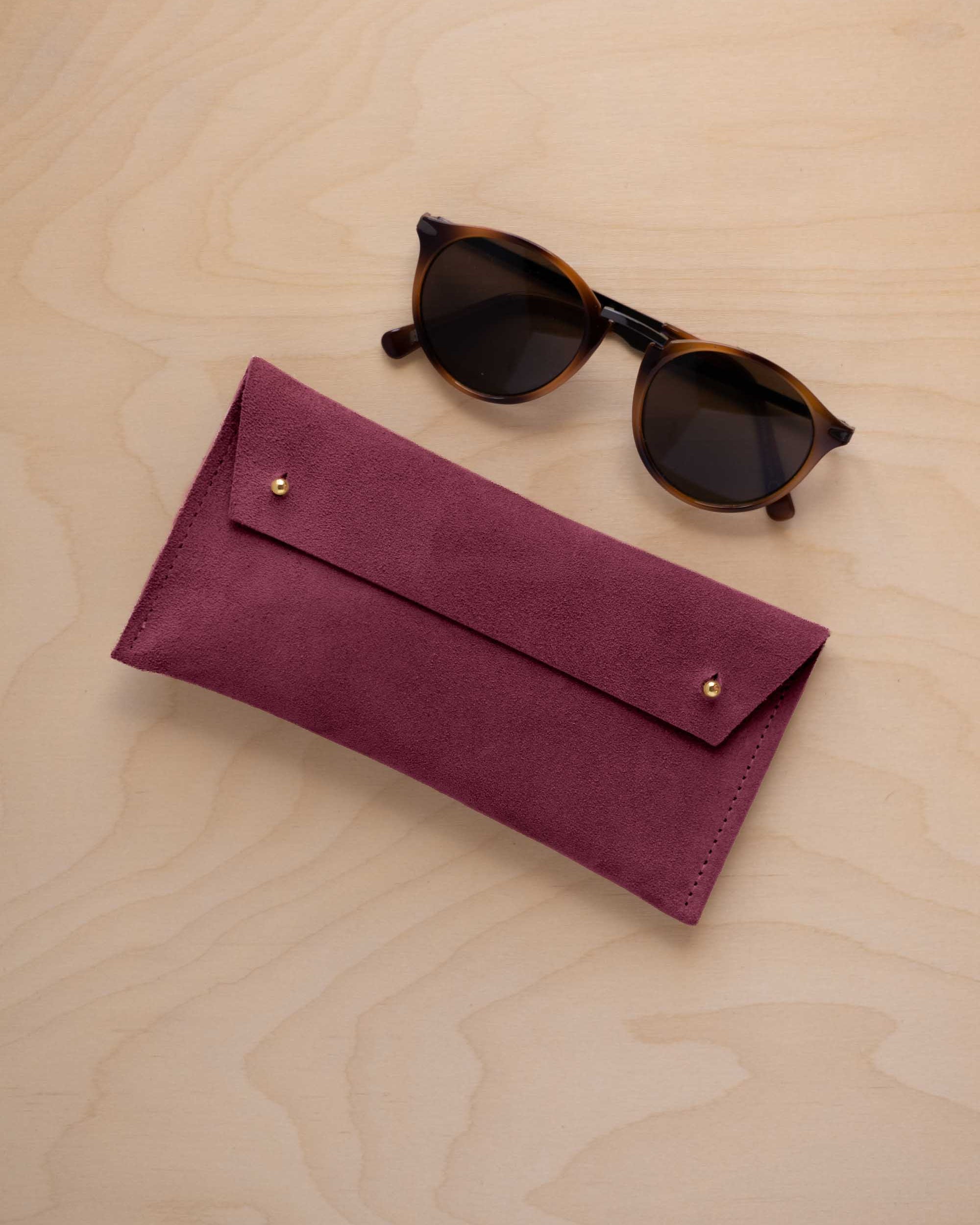 A pair of dark brown tortoiseshell sunglasses with round frames, resting on a light beige wood-grain surface next to a burgundy suede glasses case with gold coloured buttons.