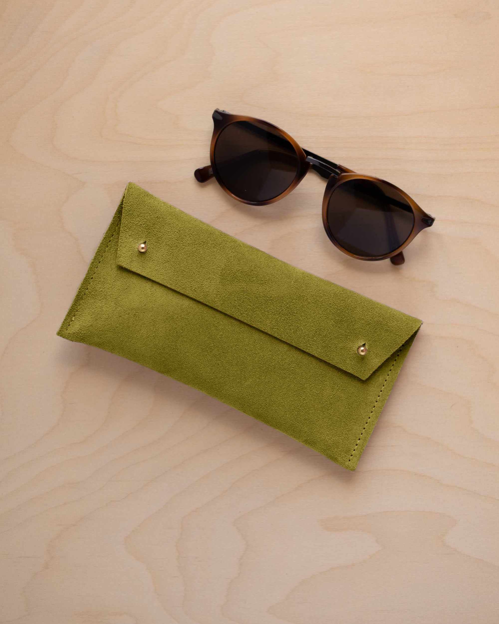 A pair of dark brown tortoiseshell sunglasses with round frames, resting on a light beige wood-grain surface next to a chartreuse lime green coloured suede glasses case with gold coloured buttons.