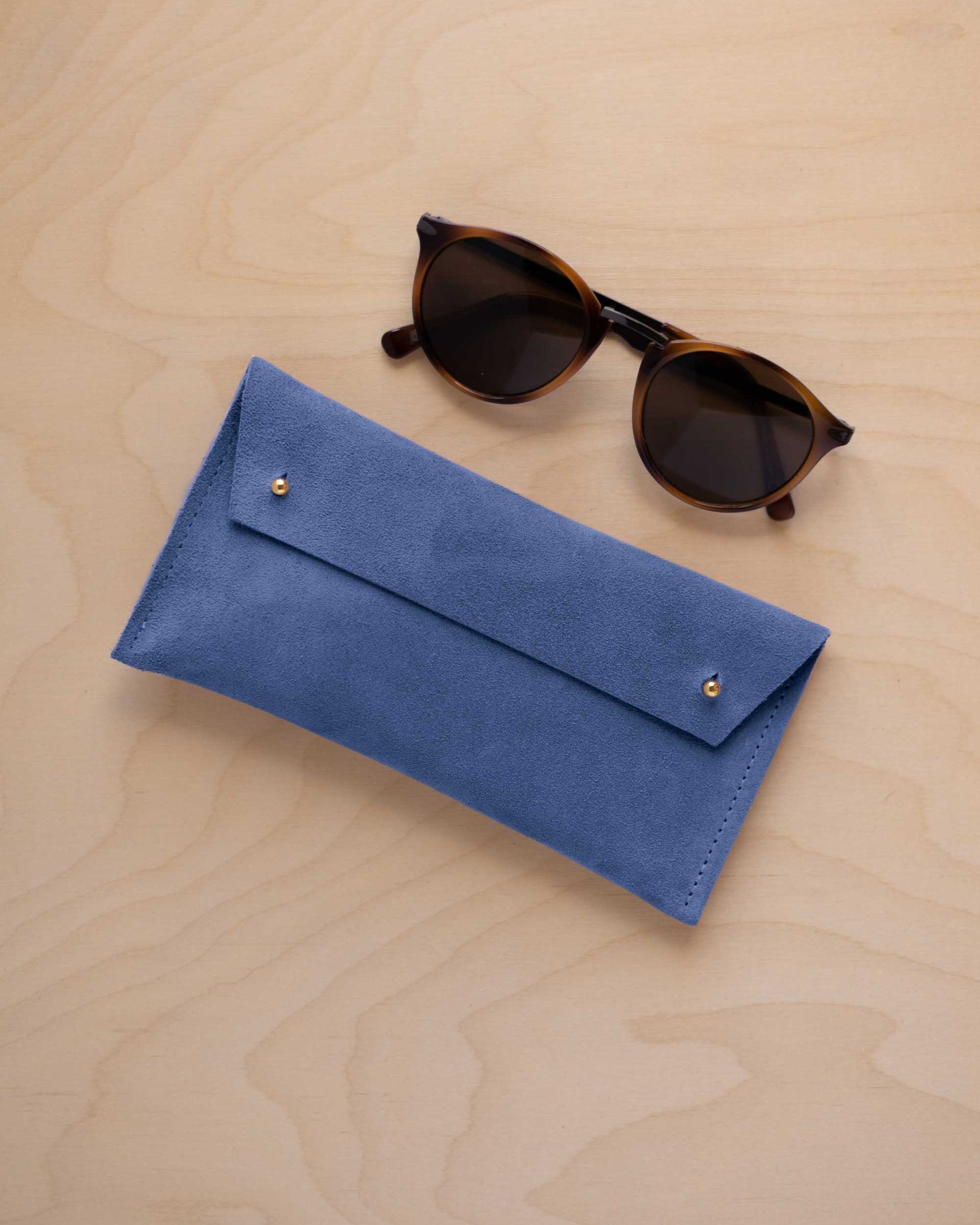 A pair of dark brown tortoiseshell sunglasses with round frames, resting on a light beige wood-grain surface next to a cobalt blue suede glasses case with gold coloured buttons.