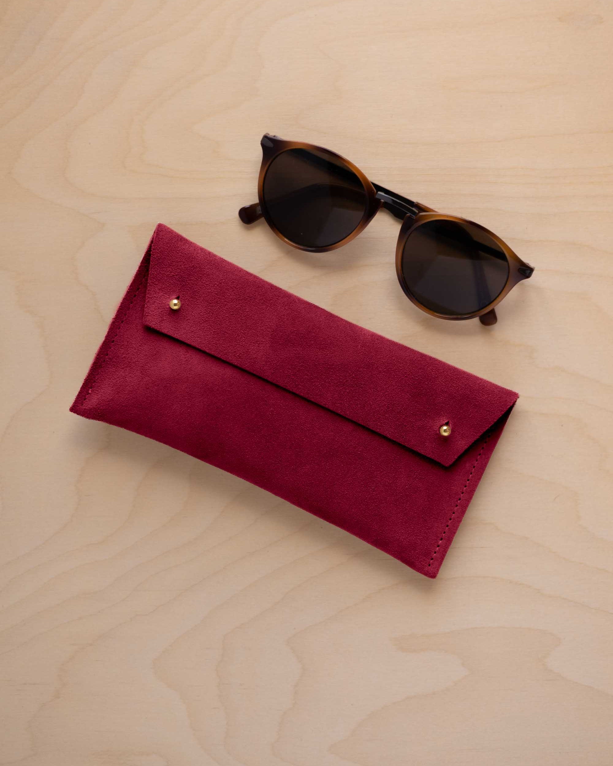 A pair of dark brown tortoiseshell sunglasses with round frames, resting on a light beige wood-grain surface next to a crimson red suede glasses case with gold coloured buttons.
