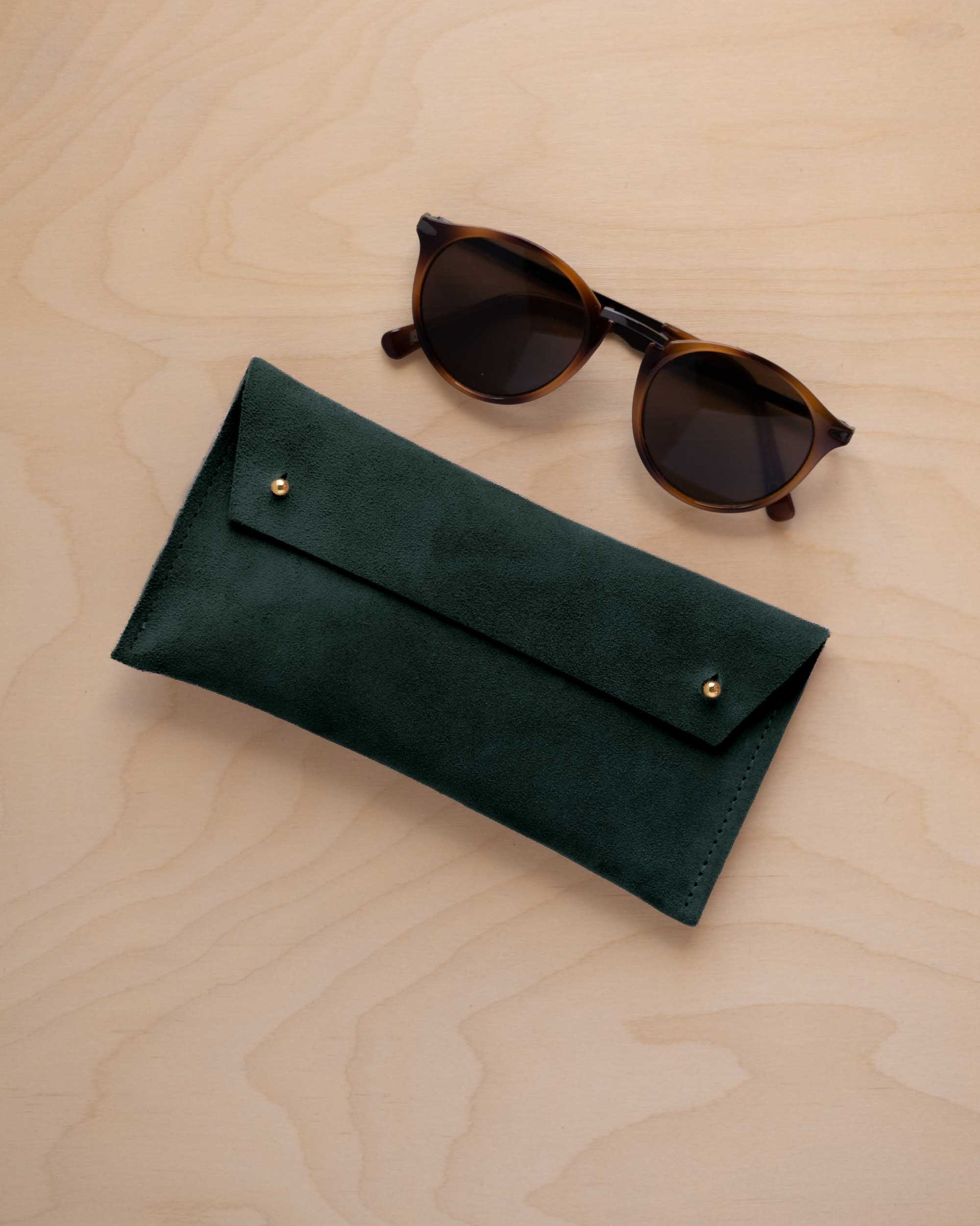 A pair of dark brown tortoiseshell sunglasses with round frames, resting on a light beige wood-grain surface next to a deep forest green suede glasses case with gold coloured buttons.
