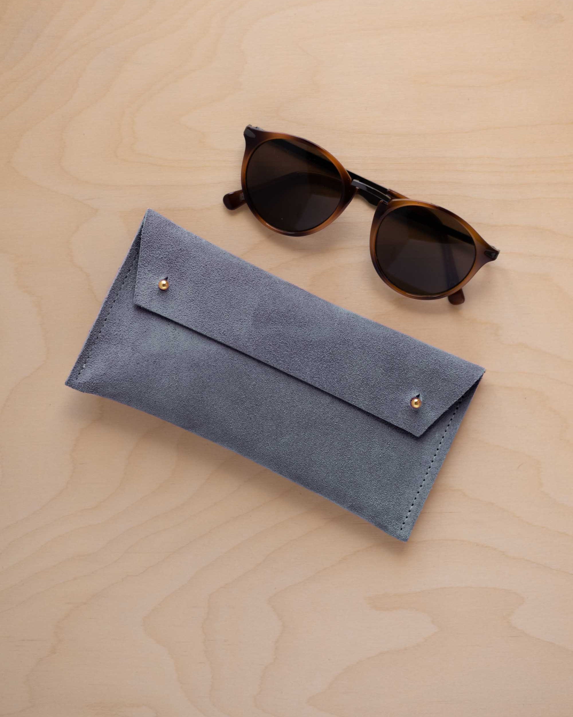 A pair of dark brown tortoiseshell sunglasses with round frames, resting on a light beige wood-grain surface next to a dusky blue suede glasses case with gold coloured buttons.