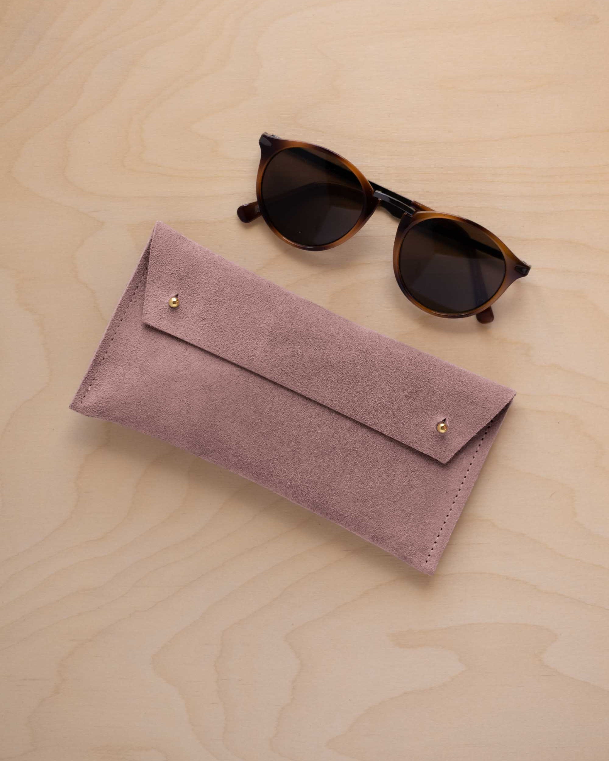 A pair of dark brown tortoiseshell sunglasses with round frames, resting on a light beige wood-grain surface next to a dusty mauve grey suede glasses case with gold coloured buttons.
