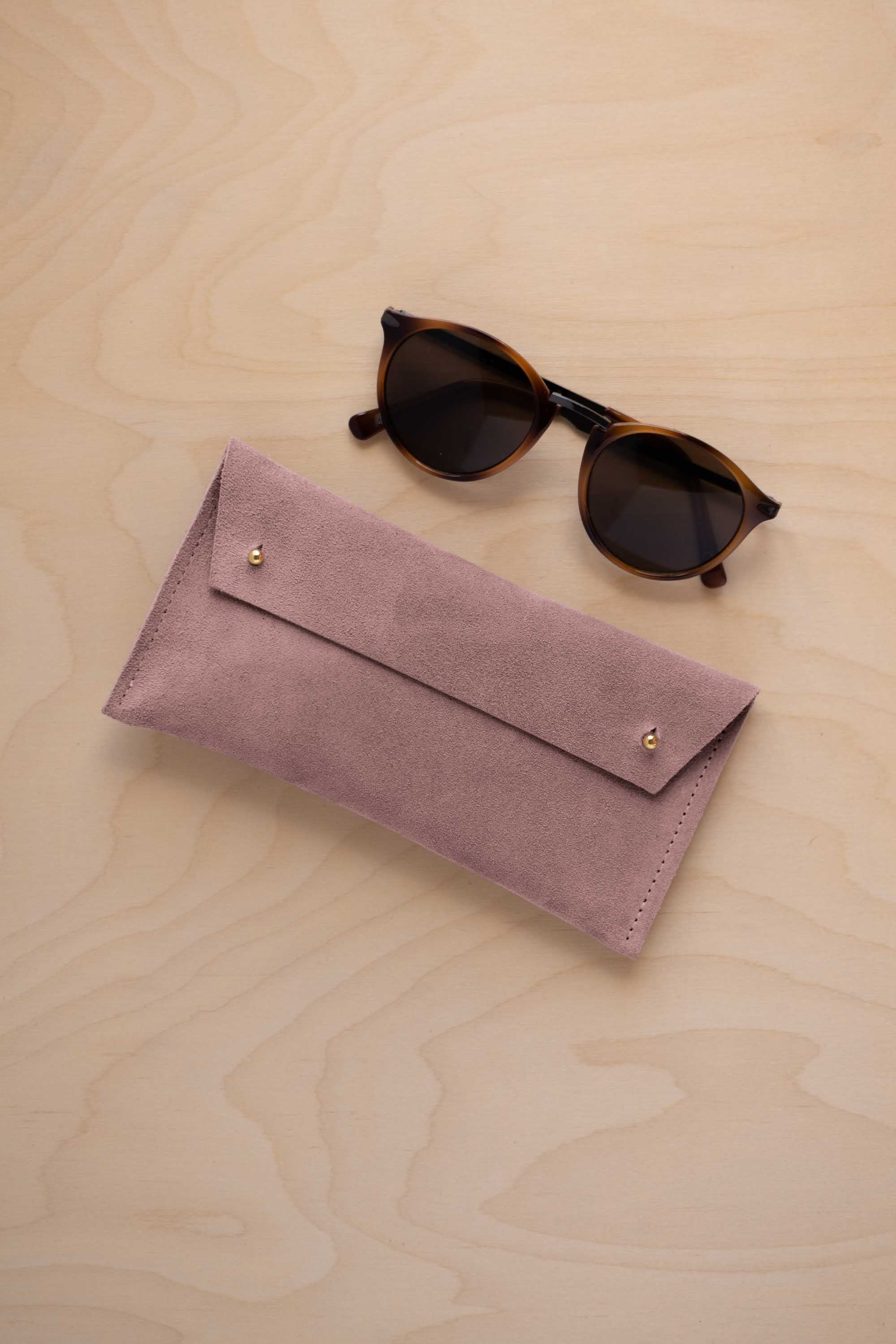 A pair of dark brown tortoiseshell sunglasses with round frames, resting on a light beige wood-grain surface next to a dusty mauve grey suede glasses case with gold coloured buttons.