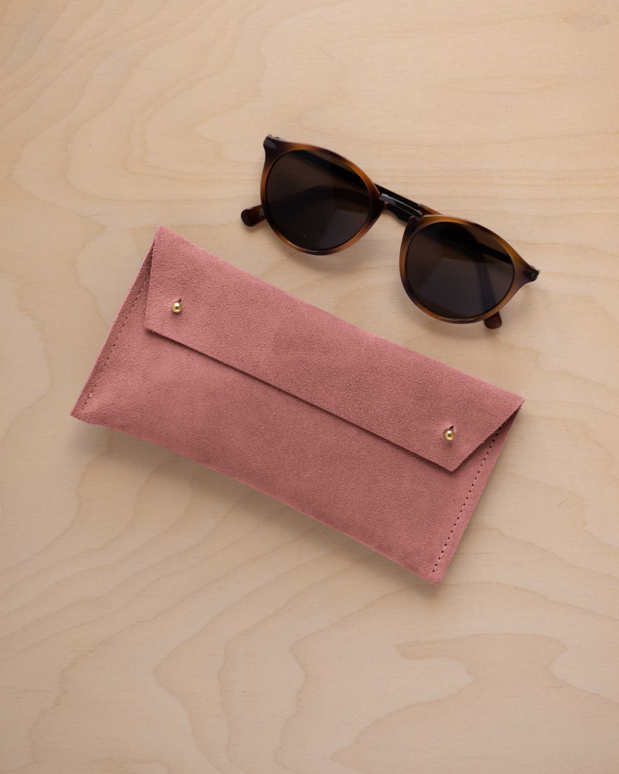 A pair of dark brown tortoiseshell sunglasses with round frames, resting on a light beige wood-grain surface next to a dusty rose pink coloured suede glasses case with gold coloured buttons.