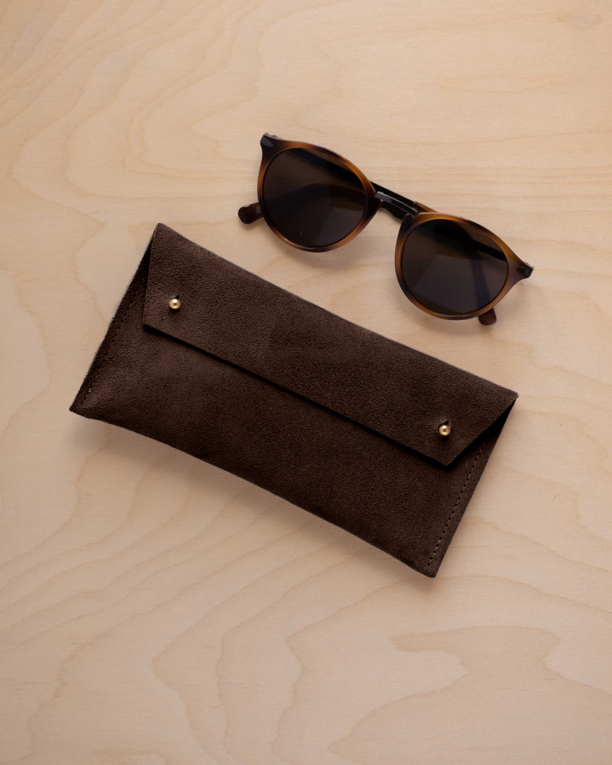 A pair of dark brown tortoiseshell sunglasses with round frames, resting on a light beige wood-grain surface next to an espresso brown coloured suede glasses case with gold coloured buttons.