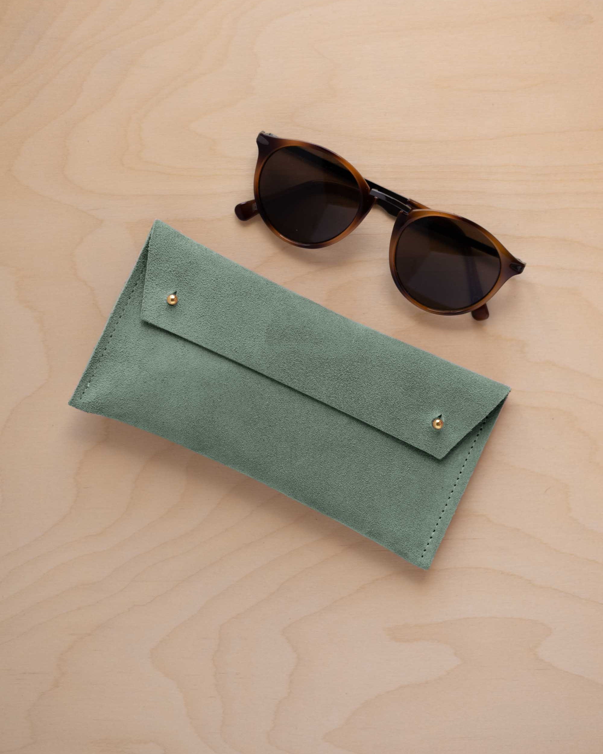 A pair of dark brown tortoiseshell sunglasses with round frames, resting on a light beige wood-grain surface next to a eucalyptus sage green coloured suede glasses case with gold coloured buttons.