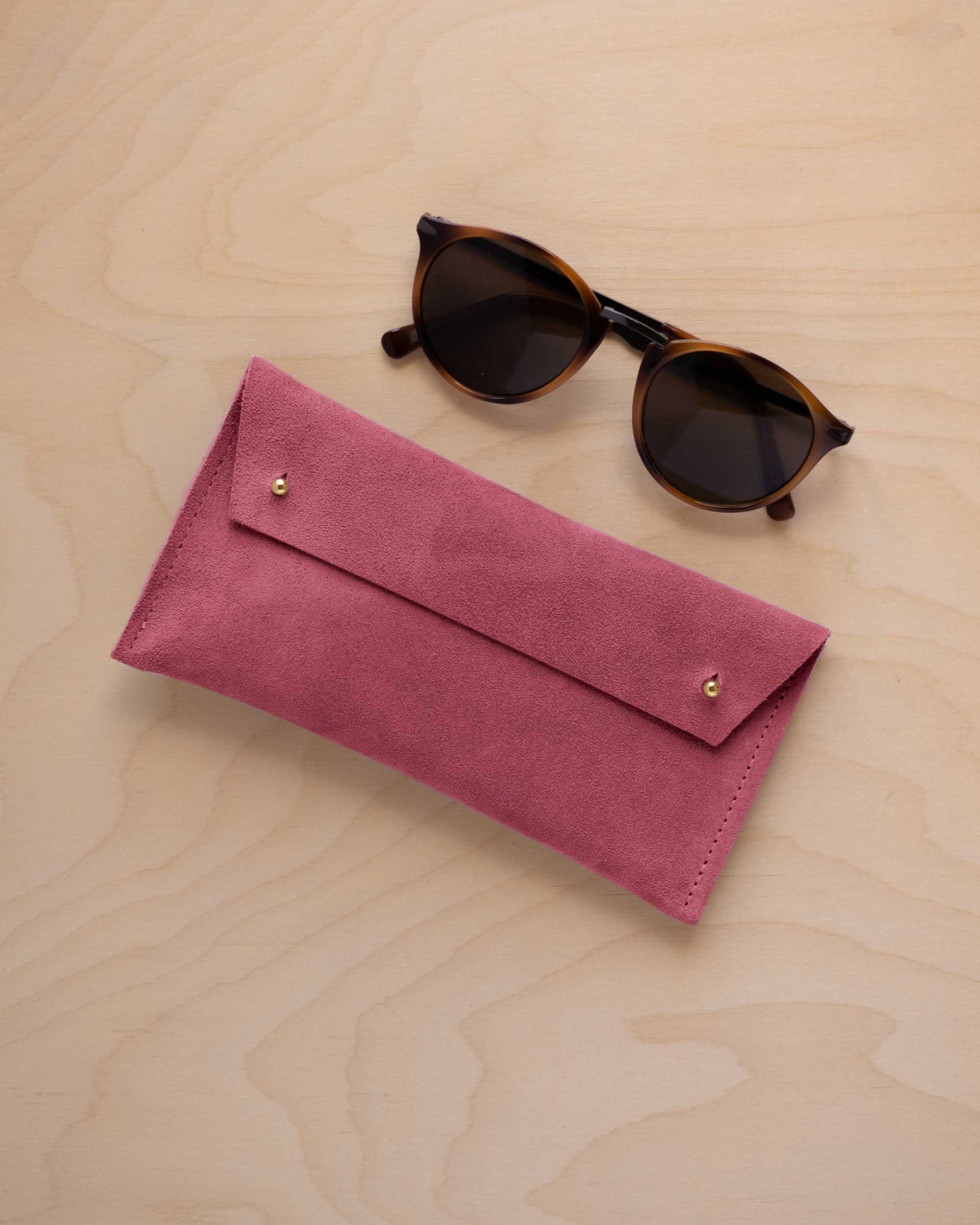 A pair of dark brown tortoiseshell sunglasses with round frames, resting on a light beige wood-grain surface next to a faded rose pink coloured suede glasses case with gold coloured buttons.