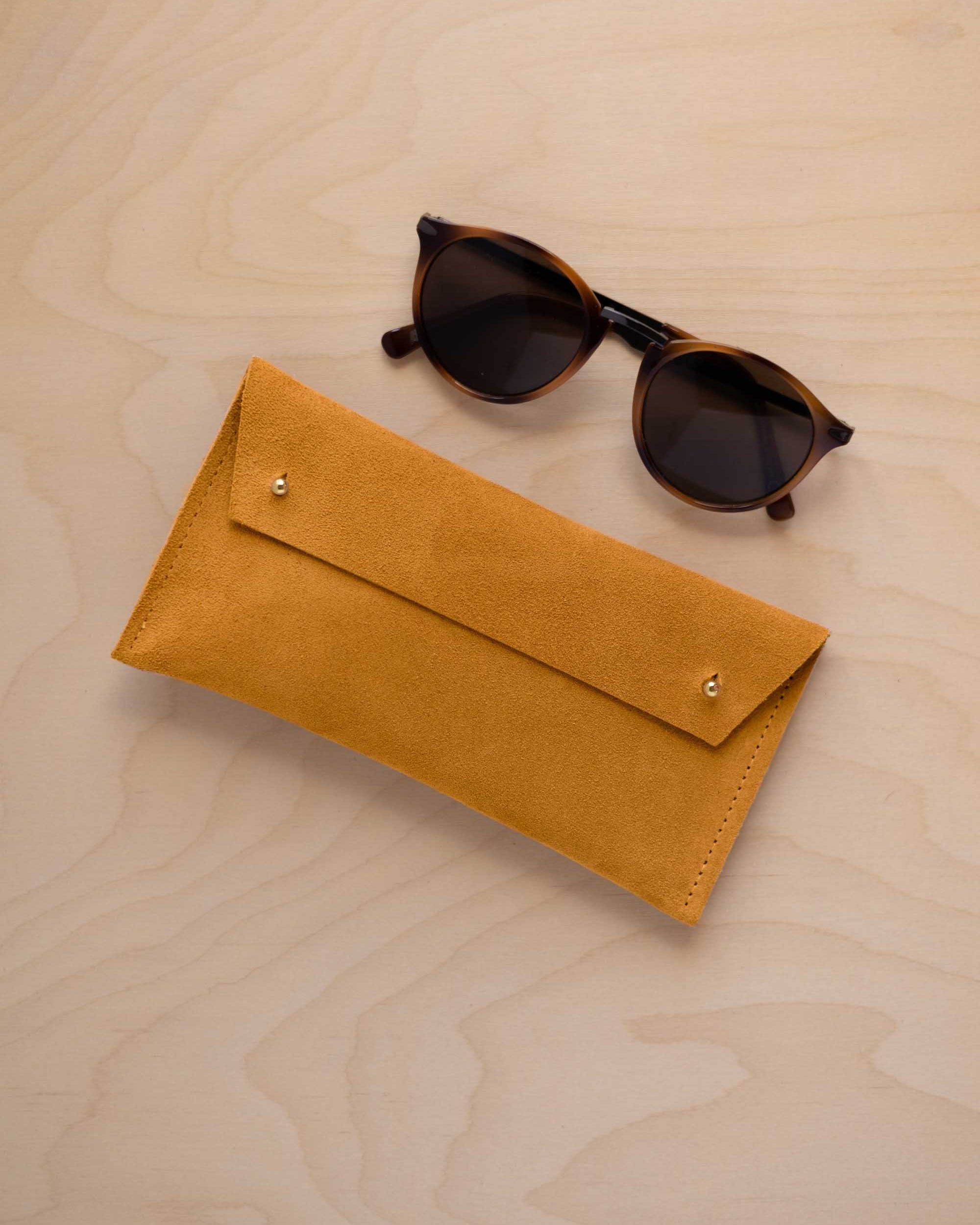 A pair of dark brown tortoiseshell sunglasses with round frames, resting on a light beige wood-grain surface next to a warm beige suede glasses case with gold coloured buttons.