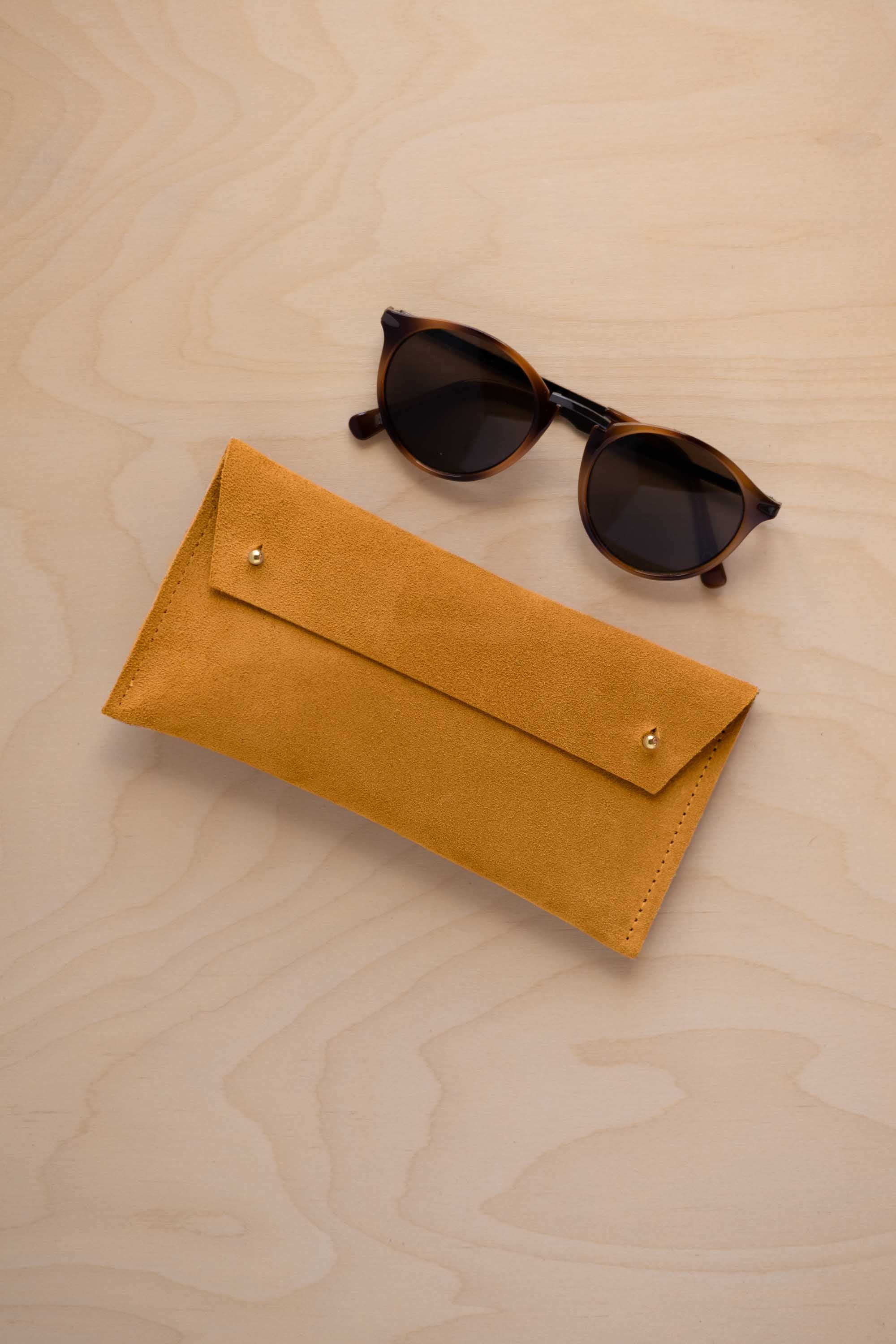 A pair of dark brown tortoiseshell sunglasses with round frames, resting on a light beige wood-grain surface next to a warm beige suede glasses case with gold coloured buttons.