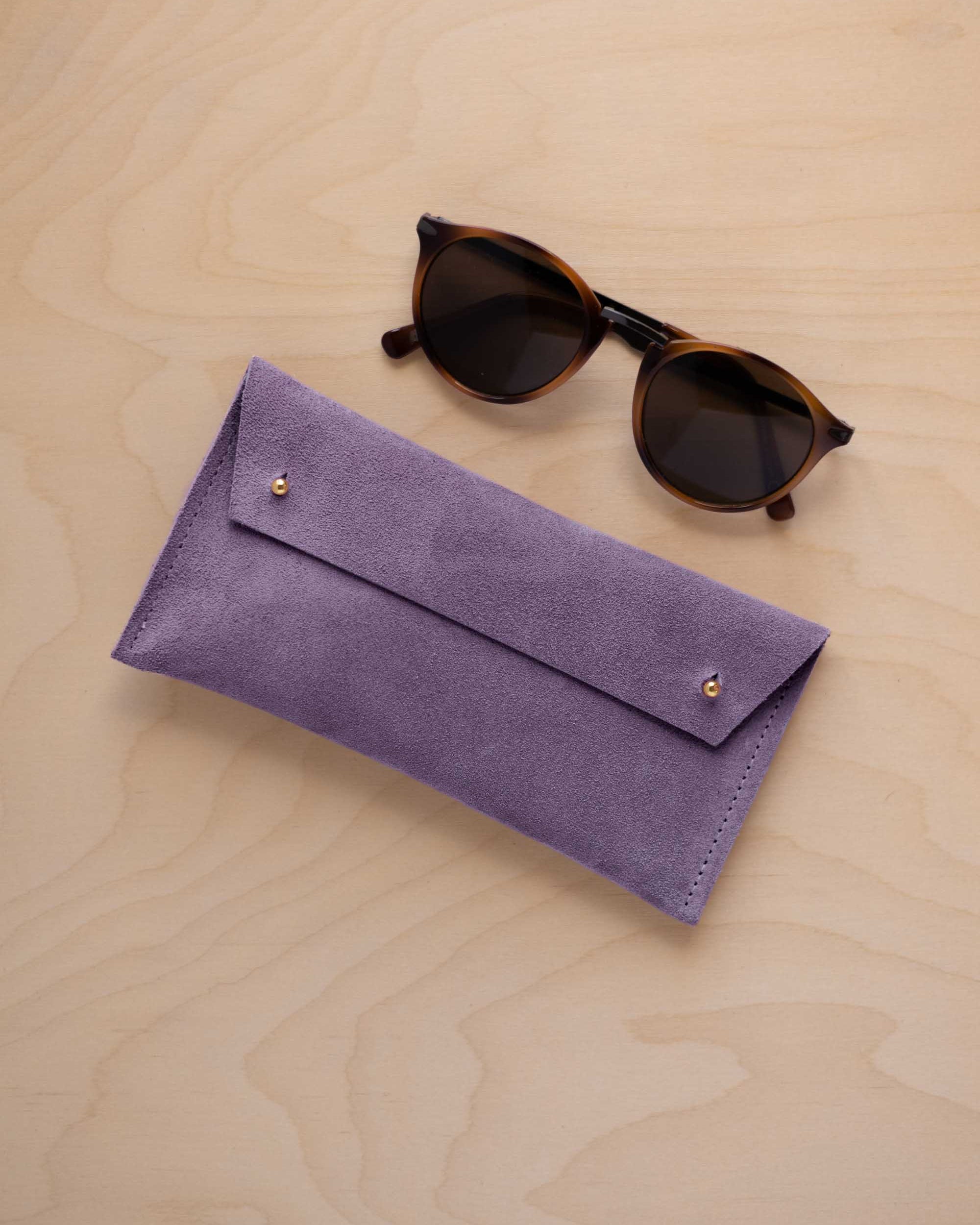 A pair of dark brown tortoiseshell sunglasses with round frames, resting on a light beige wood-grain surface next to a lavender suede glasses case with gold coloured buttons.