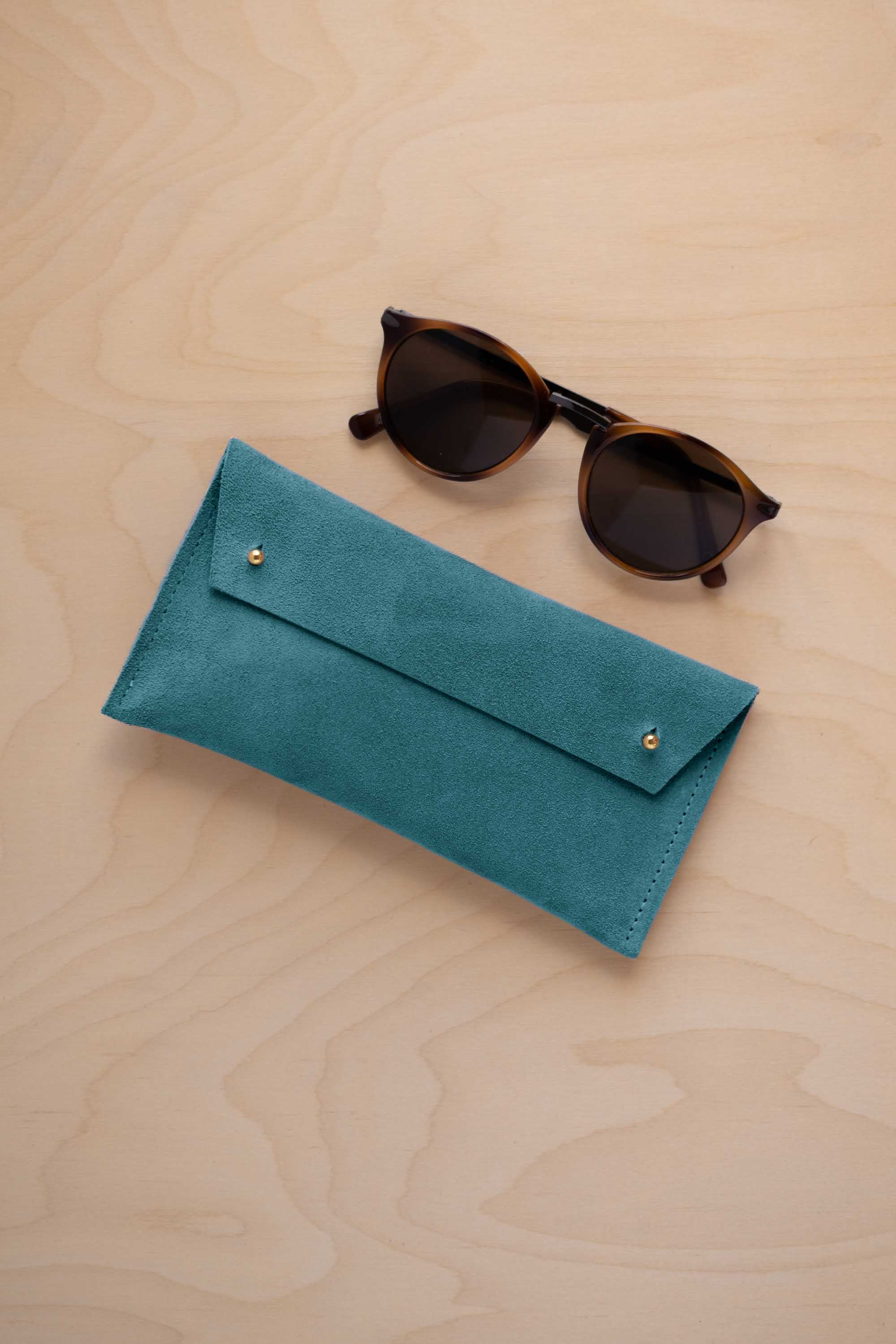A pair of dark brown tortoiseshell sunglasses with round frames, resting on a light beige wood-grain surface next to a marine teal coloured suede glasses case with gold coloured buttons.