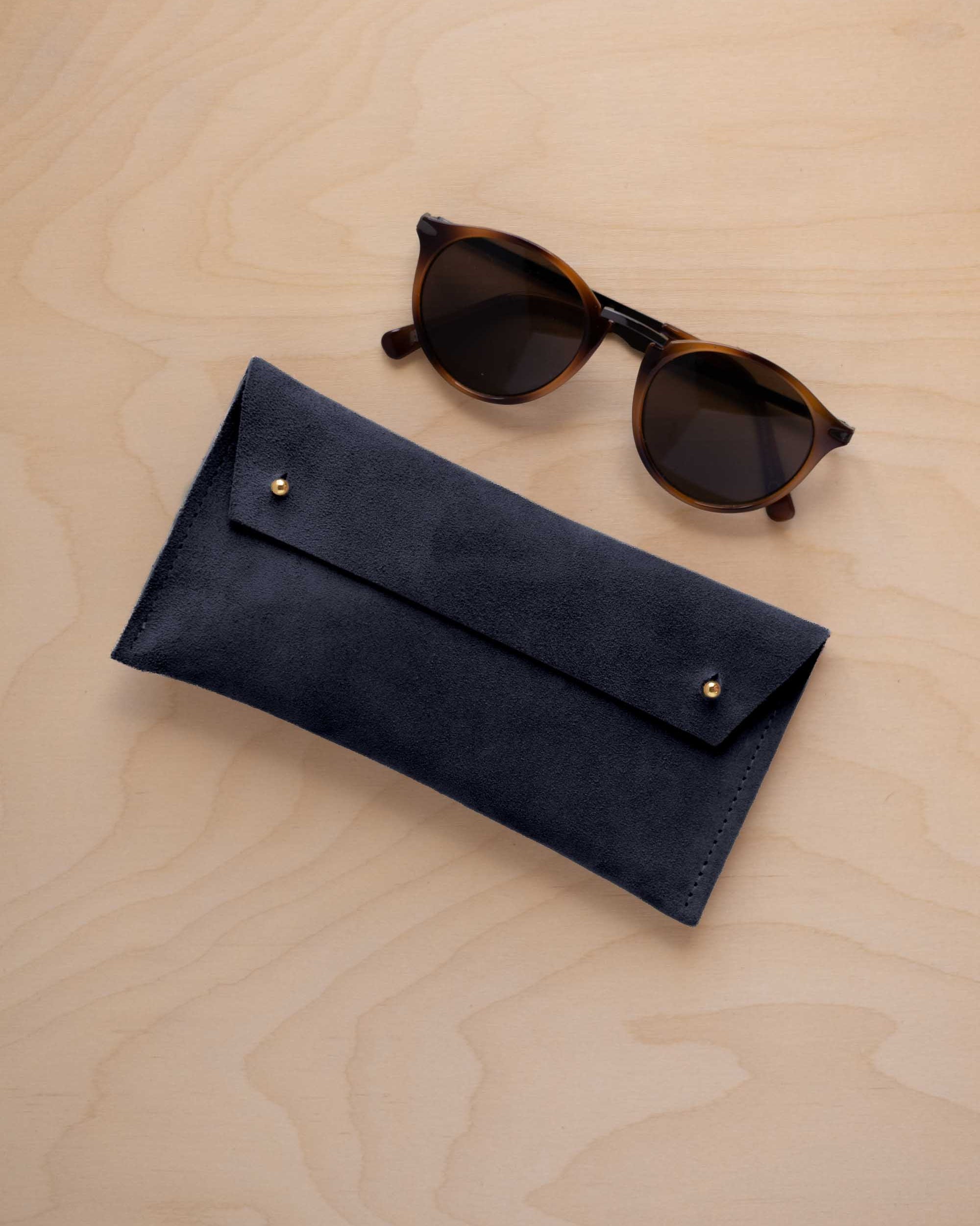 A pair of dark brown tortoiseshell sunglasses with round frames, resting on a light beige wood-grain surface next to a midnight navy blue suede glasses case with gold coloured buttons.