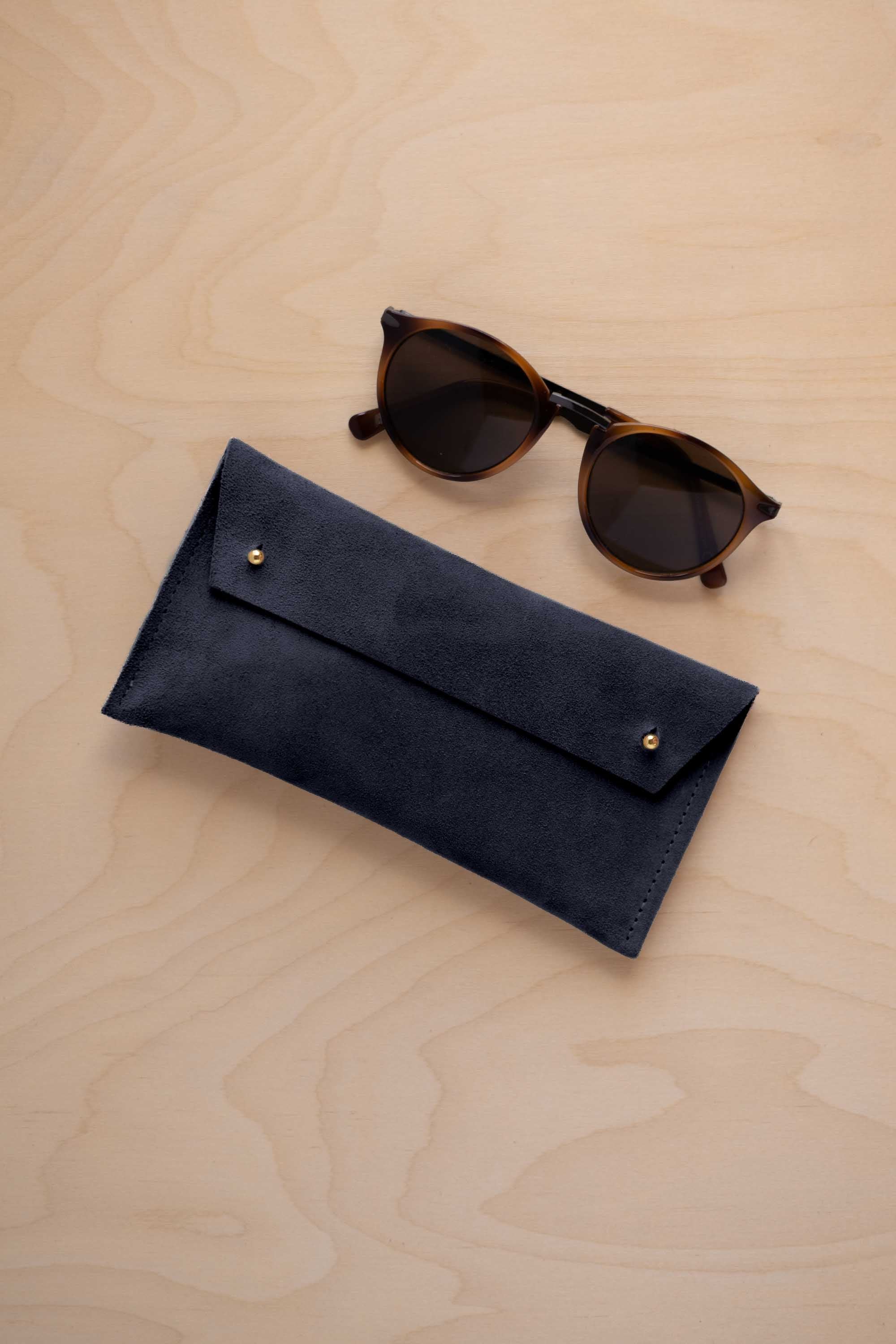 A pair of dark brown tortoiseshell sunglasses with round frames, resting on a light beige wood-grain surface next to a midnight navy blue suede glasses case with gold coloured buttons.