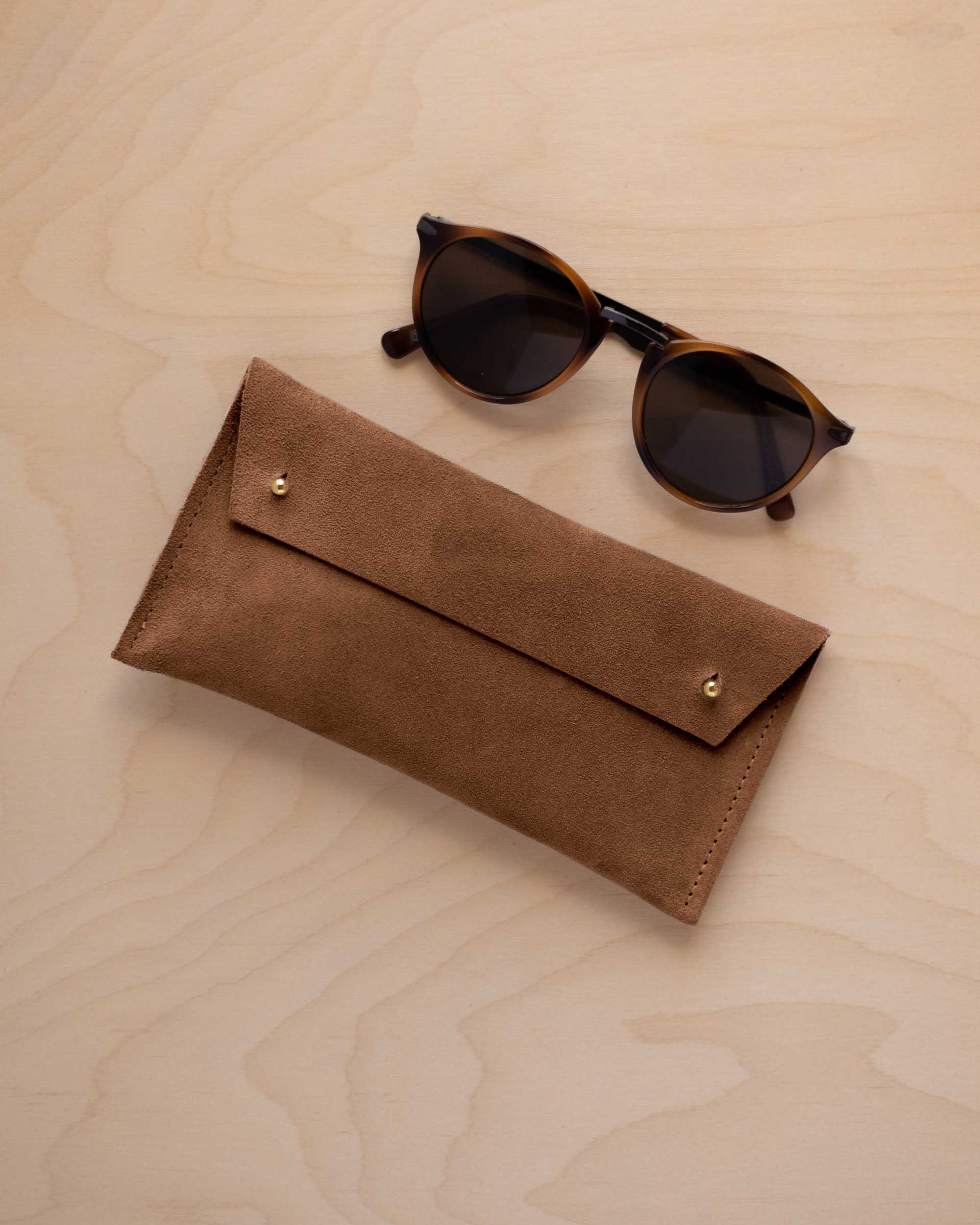 A pair of dark brown tortoiseshell sunglasses with round frames, resting on a light beige wood-grain surface next to a mocha brown suede glasses case with gold coloured buttons.