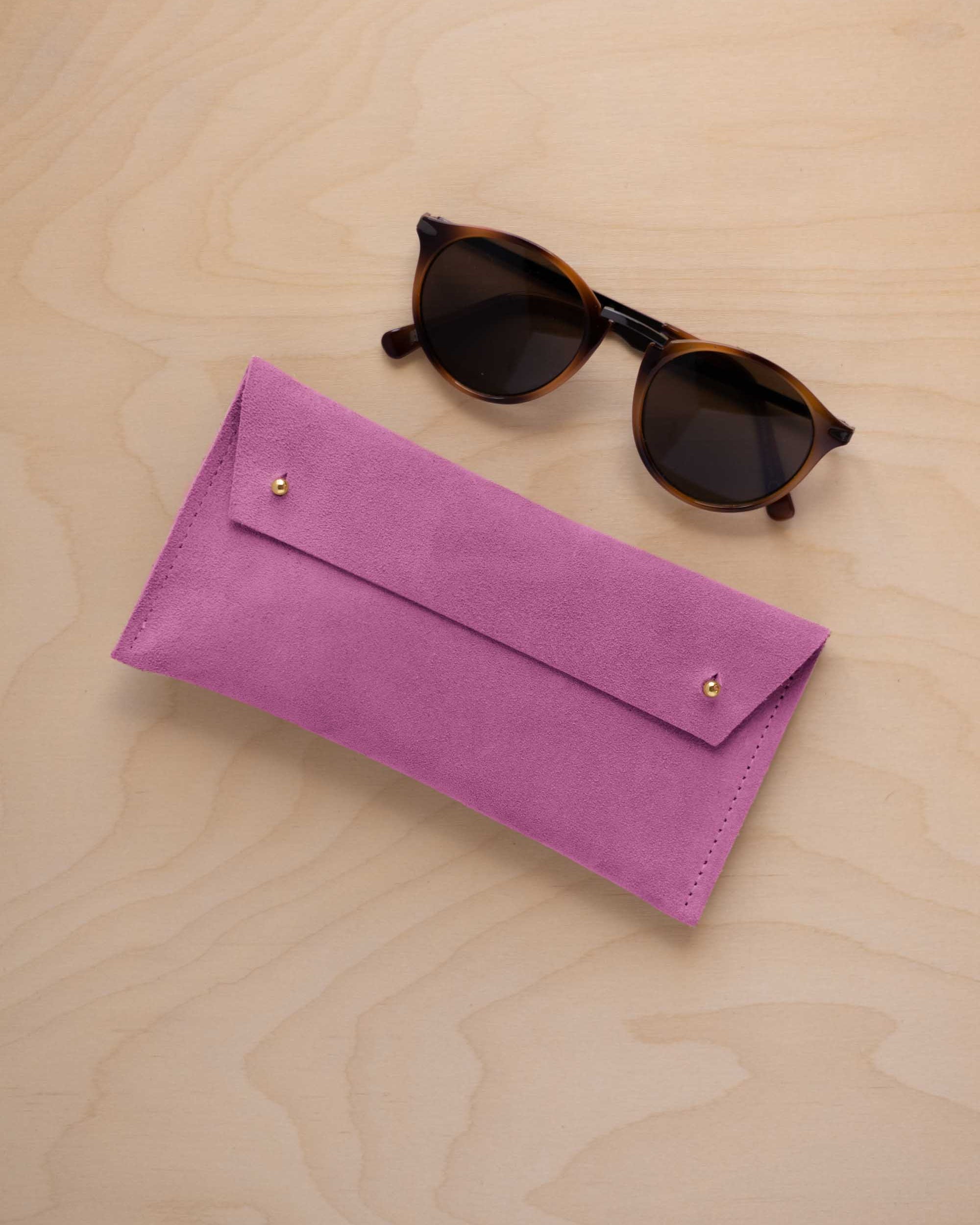 A pair of dark brown tortoiseshell sunglasses with round frames, resting on a light beige wood-grain surface next to a peony pink suede glasses case with gold coloured buttons.