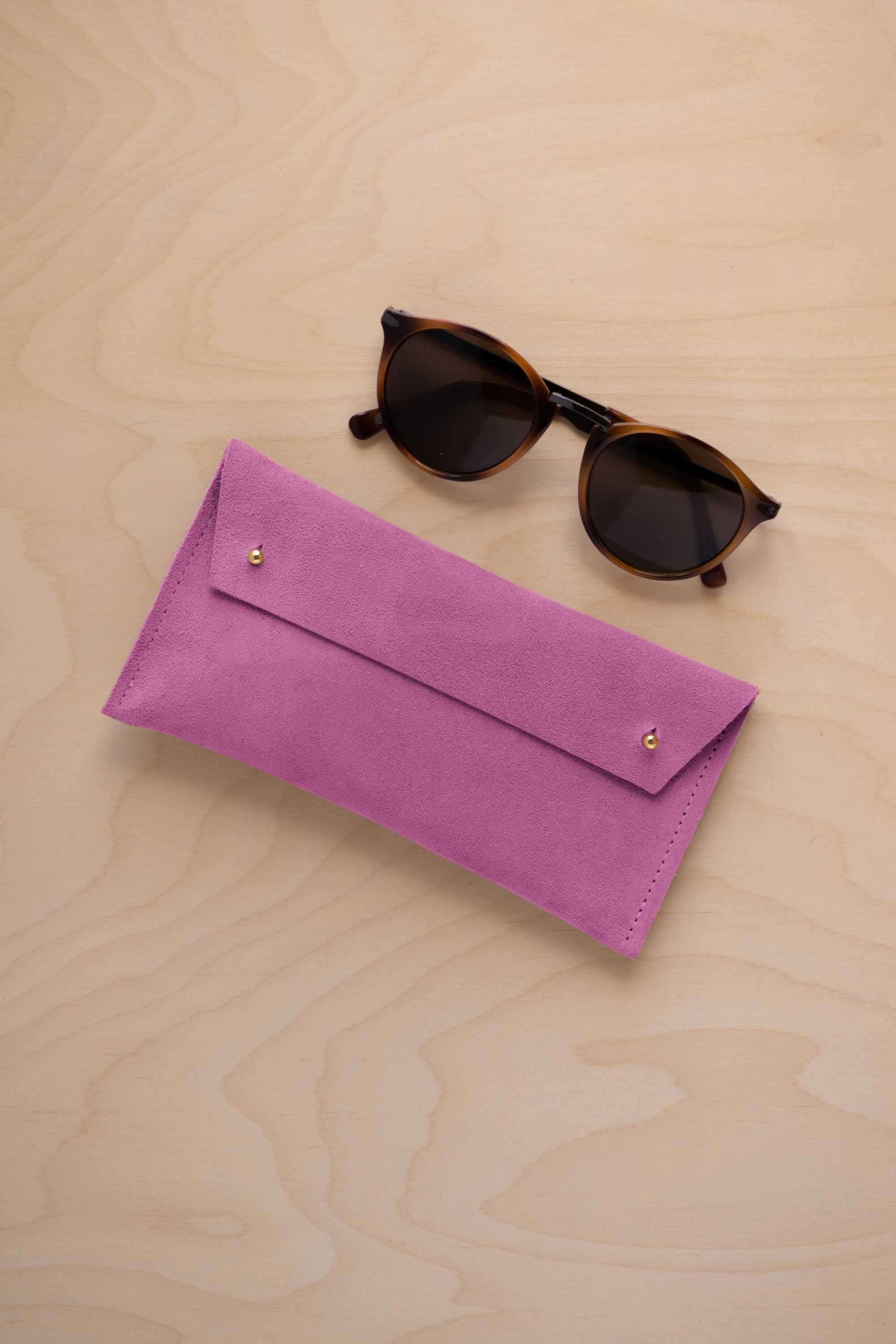 A pair of dark brown tortoiseshell sunglasses with round frames, resting on a light beige wood-grain surface next to a peony pink suede glasses case with gold coloured buttons.