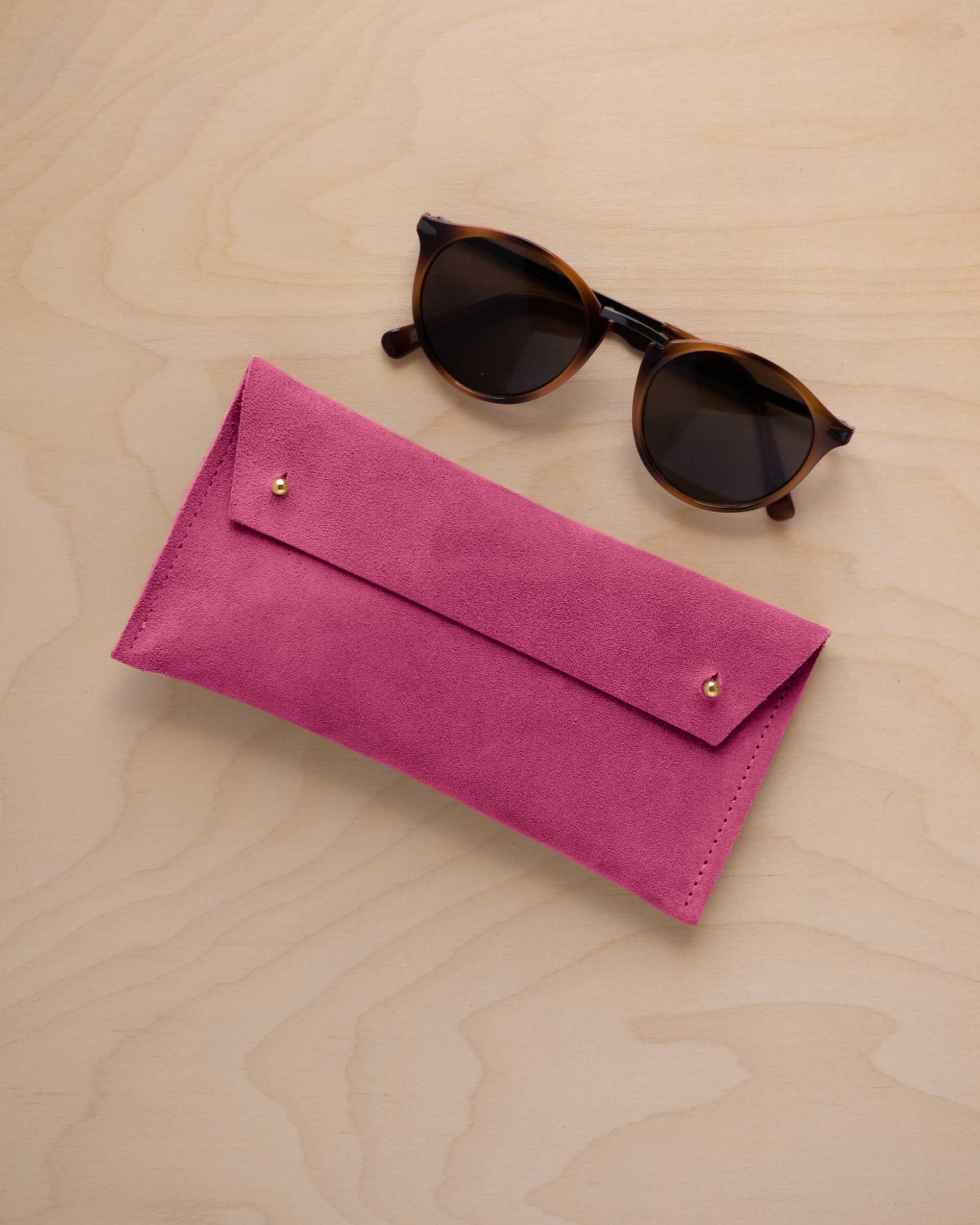 A pair of dark brown tortoiseshell sunglasses with round frames, resting on a light beige wood-grain surface next to a raspberry pink suede glasses case with gold coloured buttons.