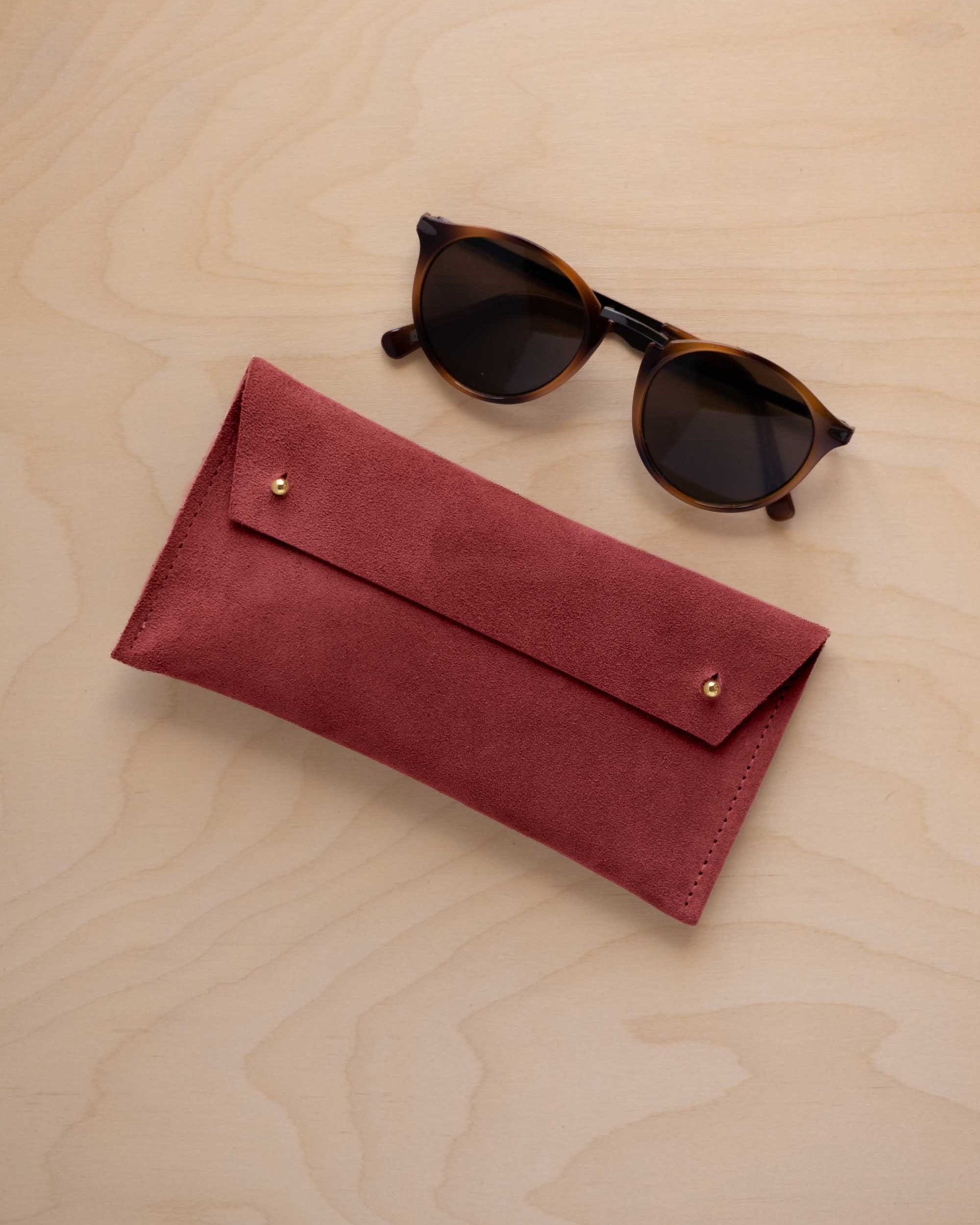 A pair of dark brown tortoiseshell sunglasses with round frames, resting on a light beige wood-grain surface next to a rust red suede glasses case with gold coloured buttons.