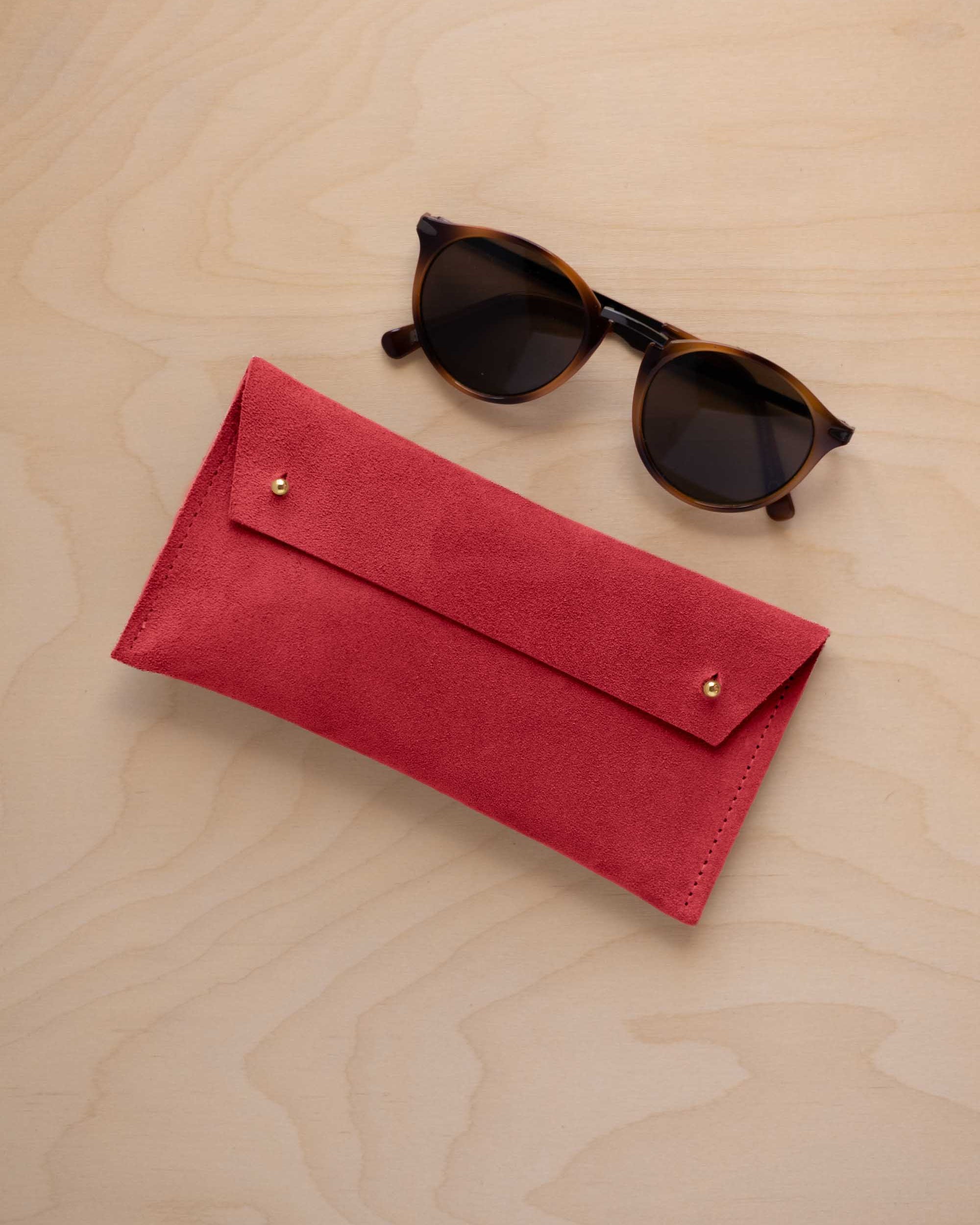 A pair of dark brown tortoiseshell sunglasses with round frames, resting on a light beige wood-grain surface next to a scarlet red suede glasses case with gold coloured buttons.