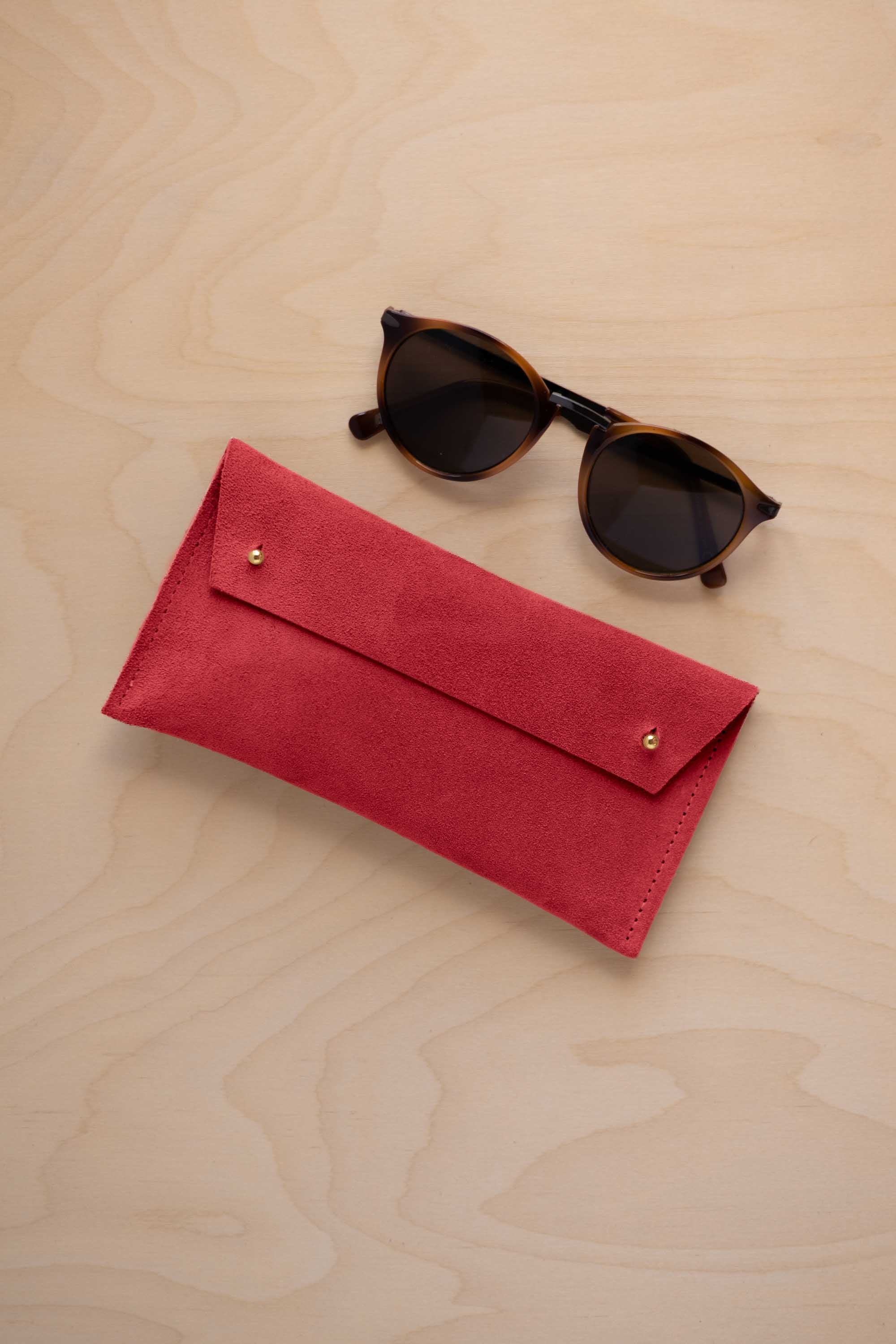 A pair of dark brown tortoiseshell sunglasses with round frames, resting on a light beige wood-grain surface next to a scarlet red suede glasses case with gold coloured buttons.
