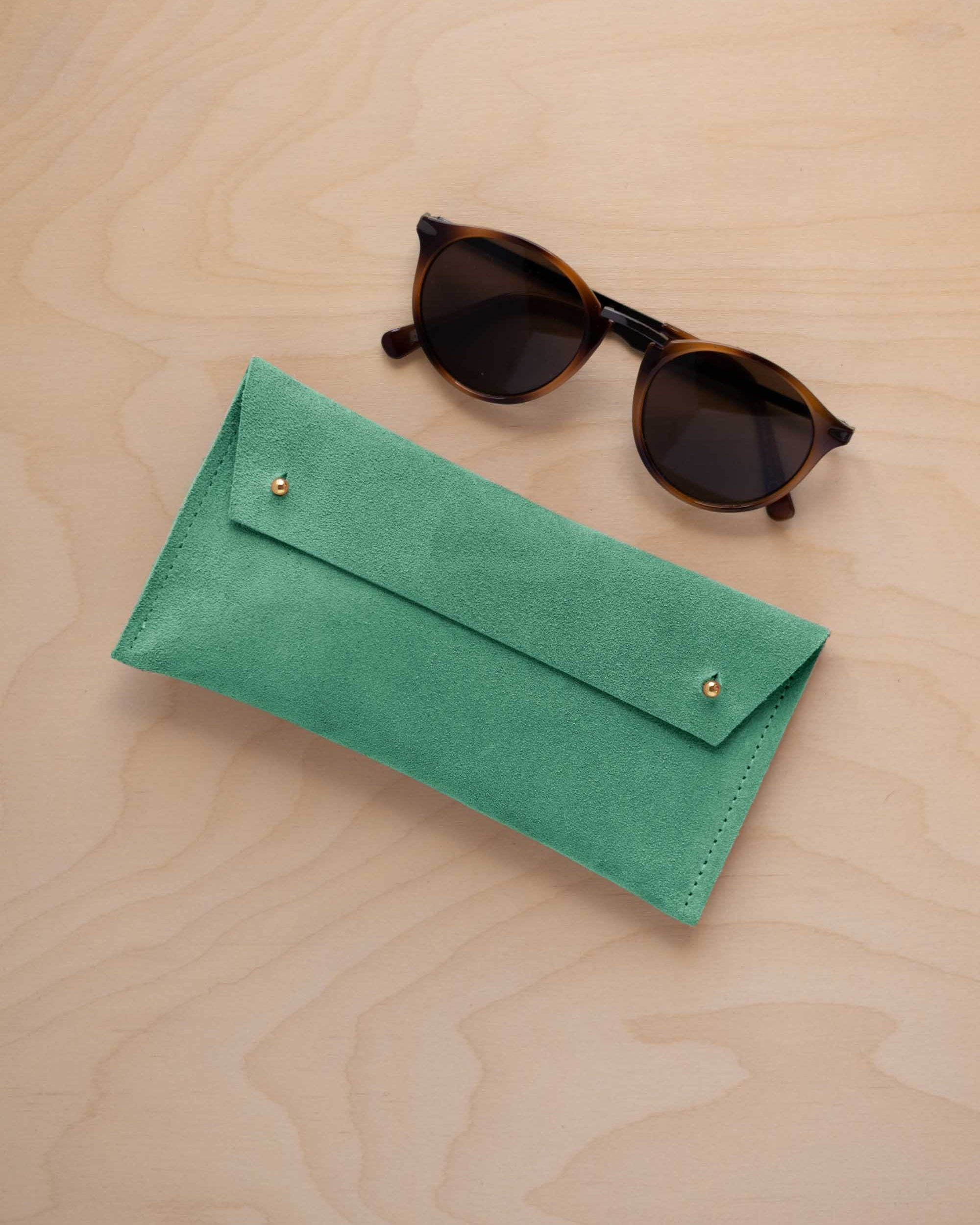 A pair of dark brown tortoiseshell sunglasses with round frames, resting on a light beige wood-grain surface next to a seafoam turquoise green coloured suede glasses case with gold coloured buttons.