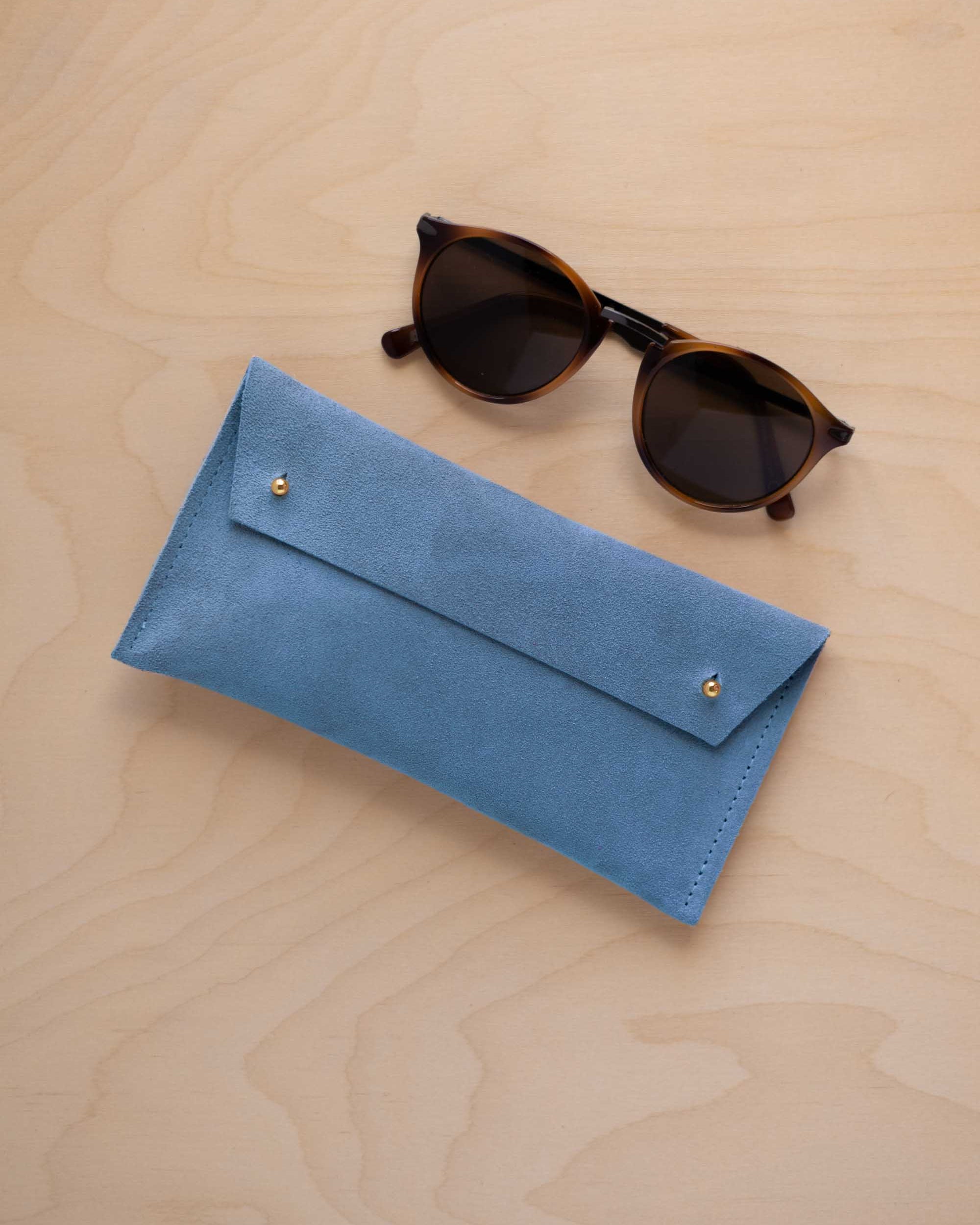 A pair of dark brown tortoiseshell sunglasses with round frames, resting on a light beige wood-grain surface next to a sky blue suede glasses case with gold coloured buttons.