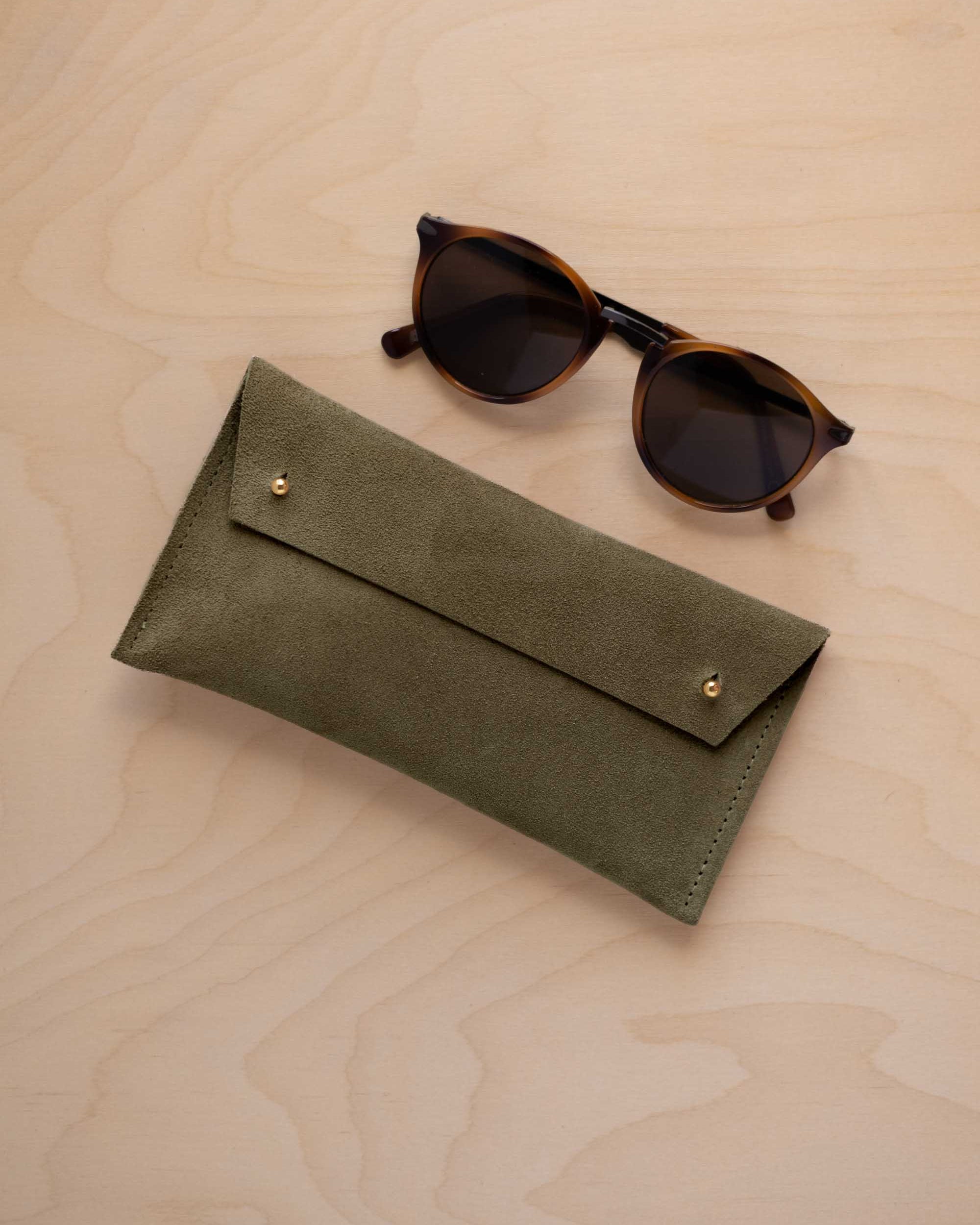 A pair of dark brown tortoiseshell sunglasses with round frames, resting on a light beige wood-grain surface next to a soft olive green coloured suede glasses case with gold coloured buttons.