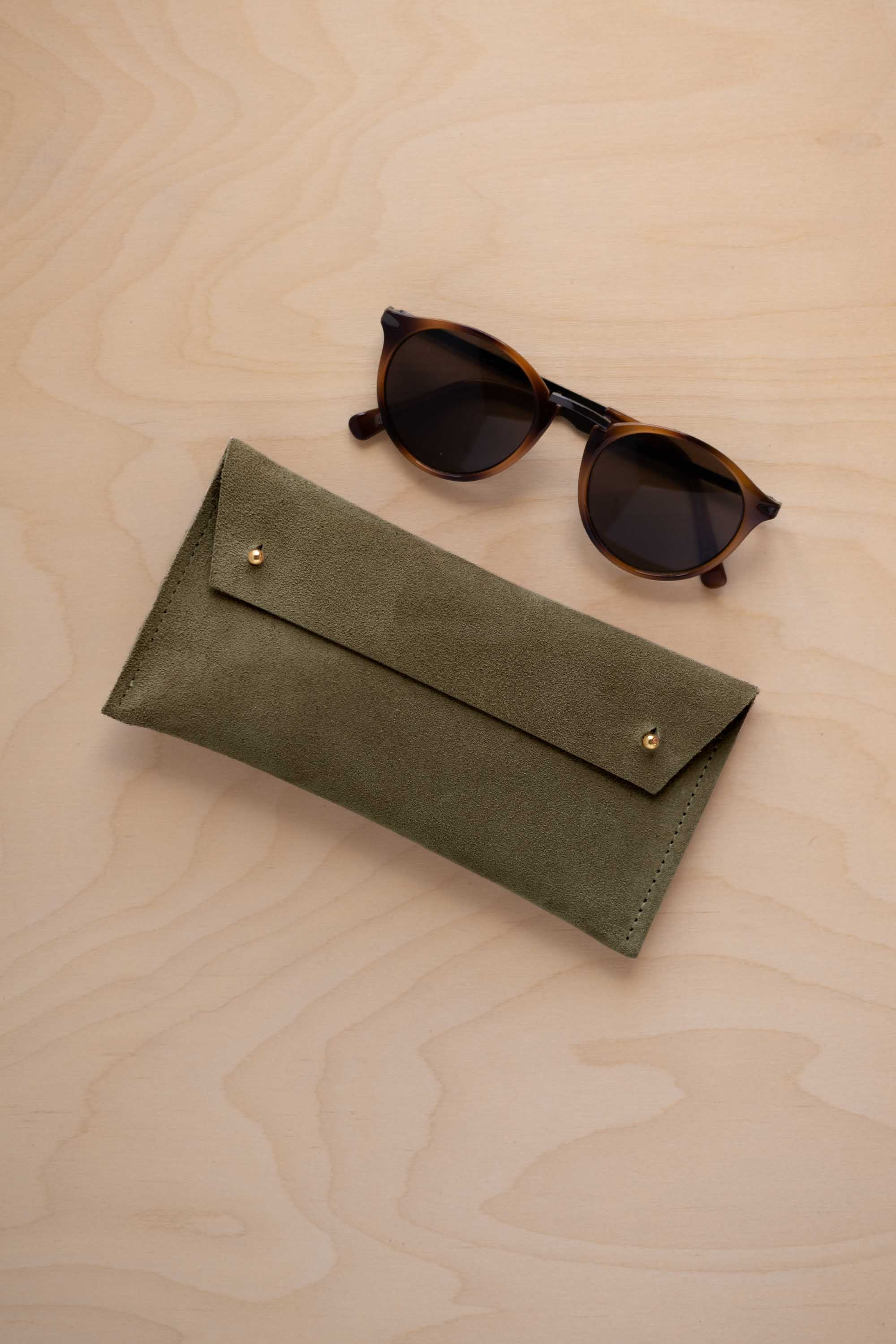 A pair of dark brown tortoiseshell sunglasses with round frames, resting on a light beige wood-grain surface next to a soft olive green coloured suede glasses case with gold coloured buttons.